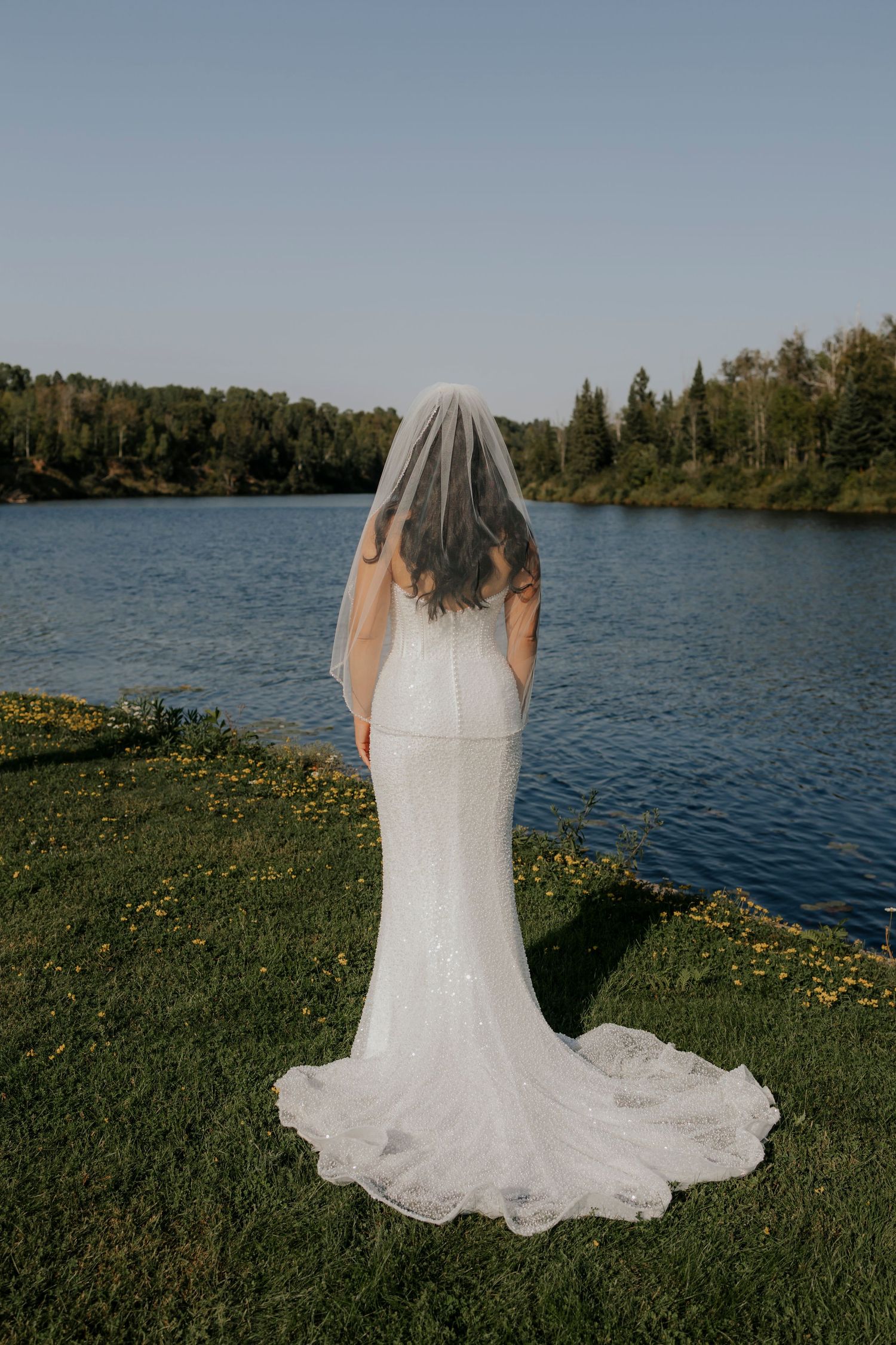 White wedding dress train spreads across green grass near a serene lake.