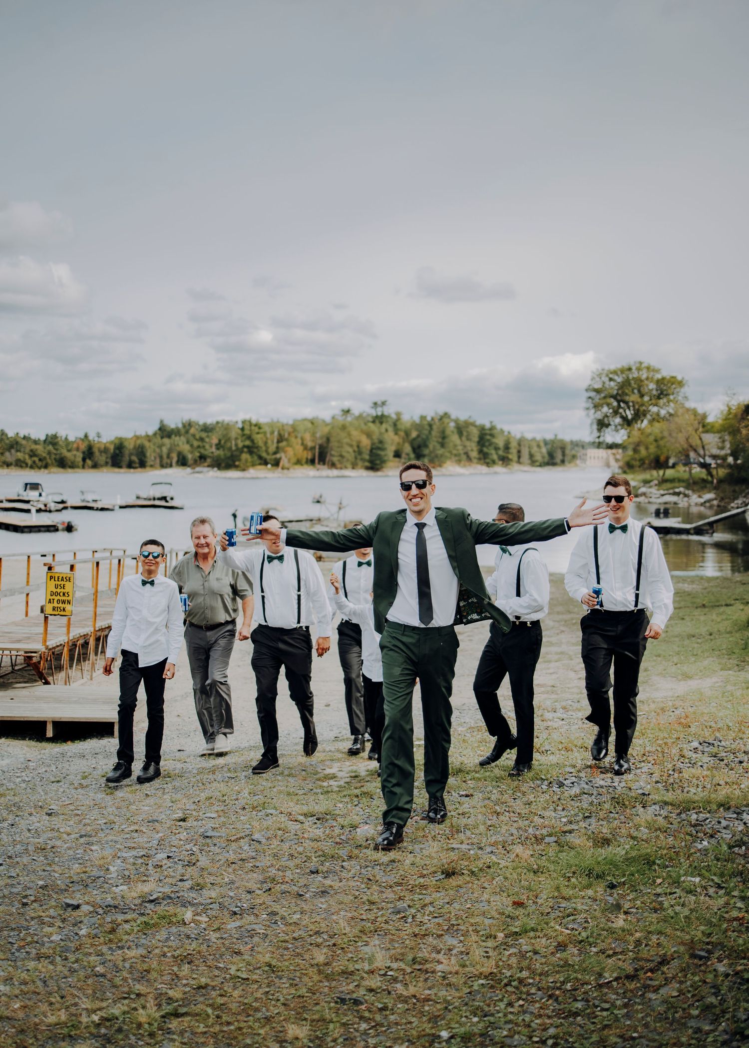 A group in formal attire stands together outdoors in a line with their arms linked near a waterfront.