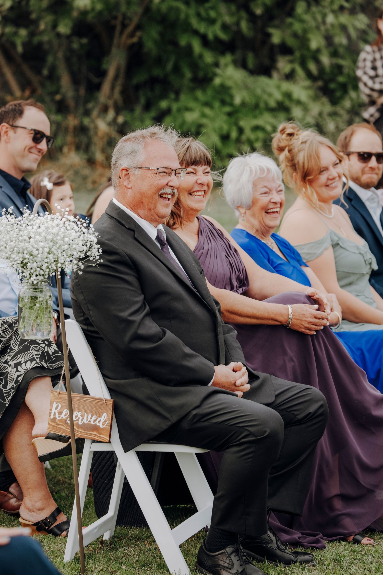 Wedding guests seated in white chairs decorated with baby's breath flowers during outdoor ceremony.