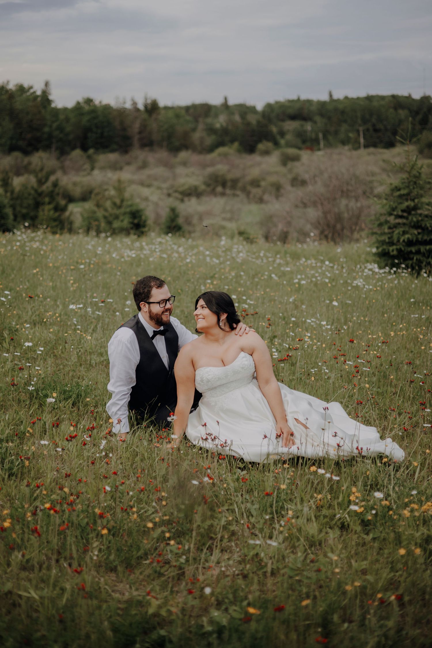 Newlyweds share an intimate moment while laying in a field of summer wildflowers under cloudy skies.