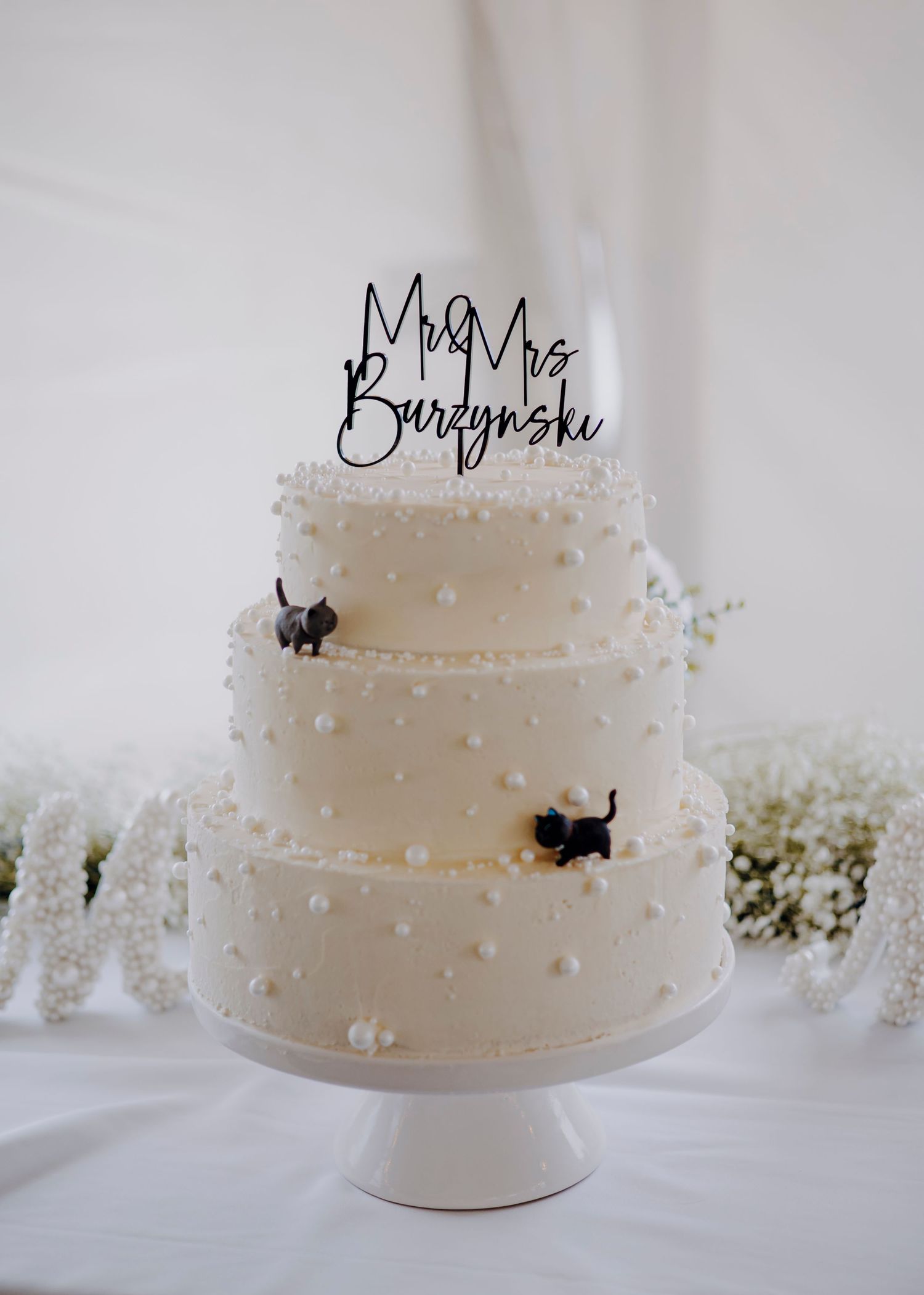 A three-tiered white wedding cake decorated with small pearls and black accents, topped with a Mr & Mrs sign.