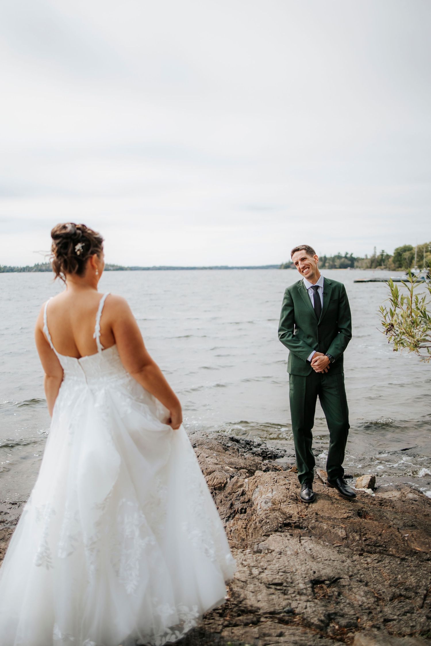Couple in wedding attire posing by rocky waterfront.