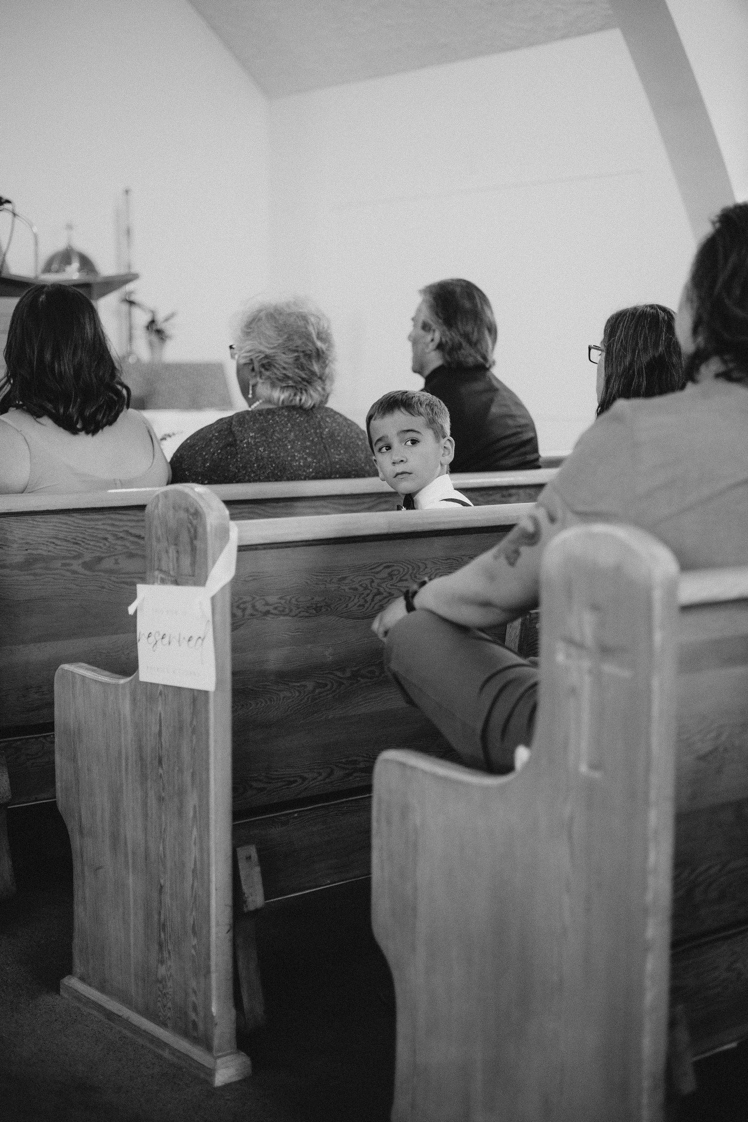 Black and white photo of wedding guests sitting in wooden church pews during ceremony.