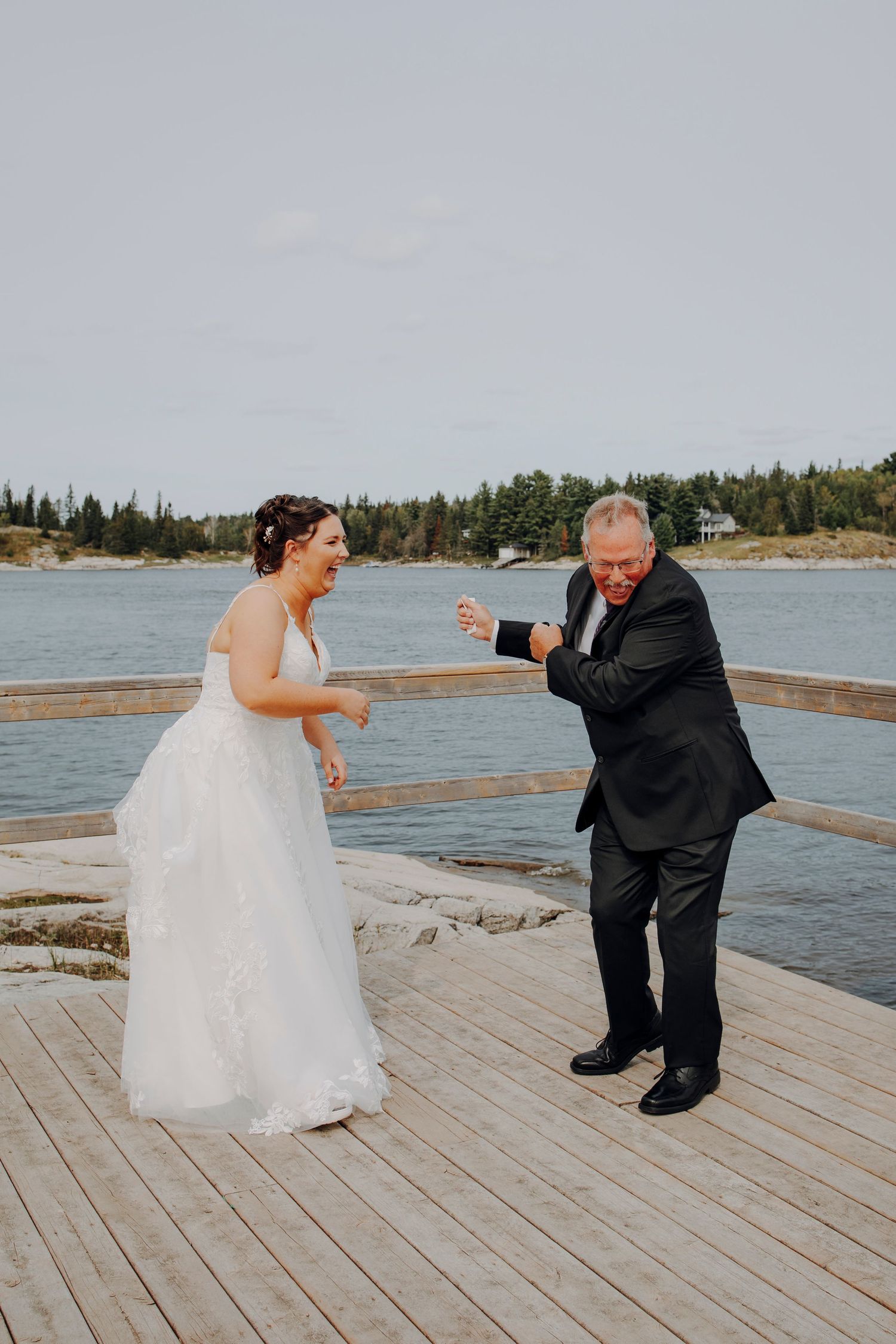 A couple dances playfully together on a wooden dock by the water.