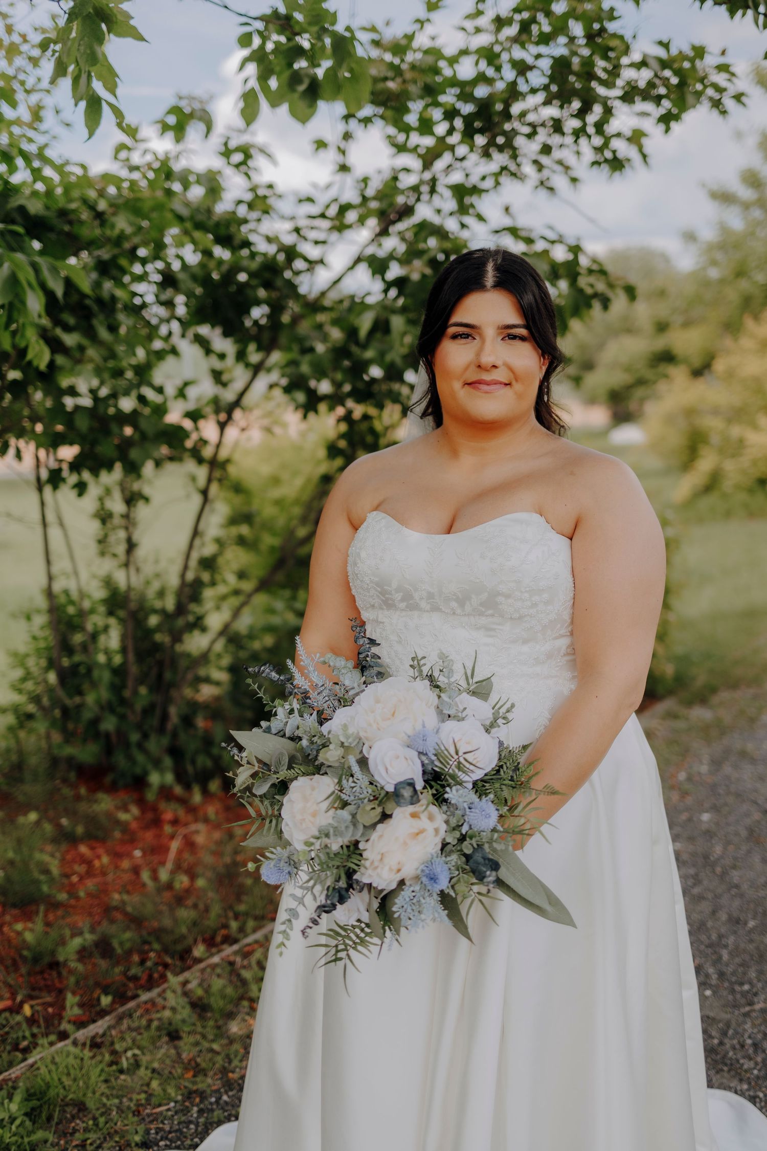 Elegant wedding bouquet featuring white and blue flowers with delicate greenery against a natural outdoor setting.
