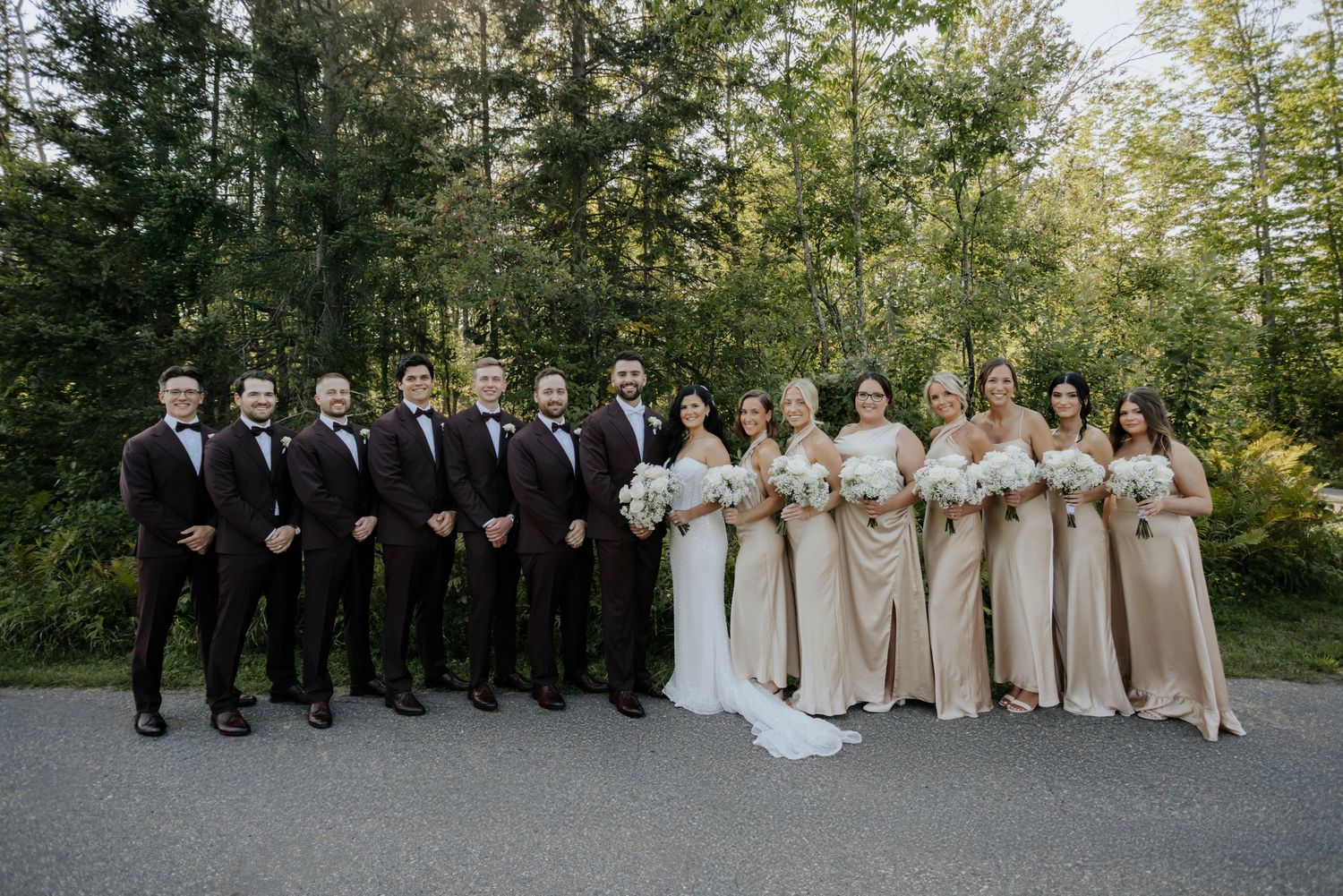Wedding party members in formal attire pose for a group photo in a wooded outdoor setting.