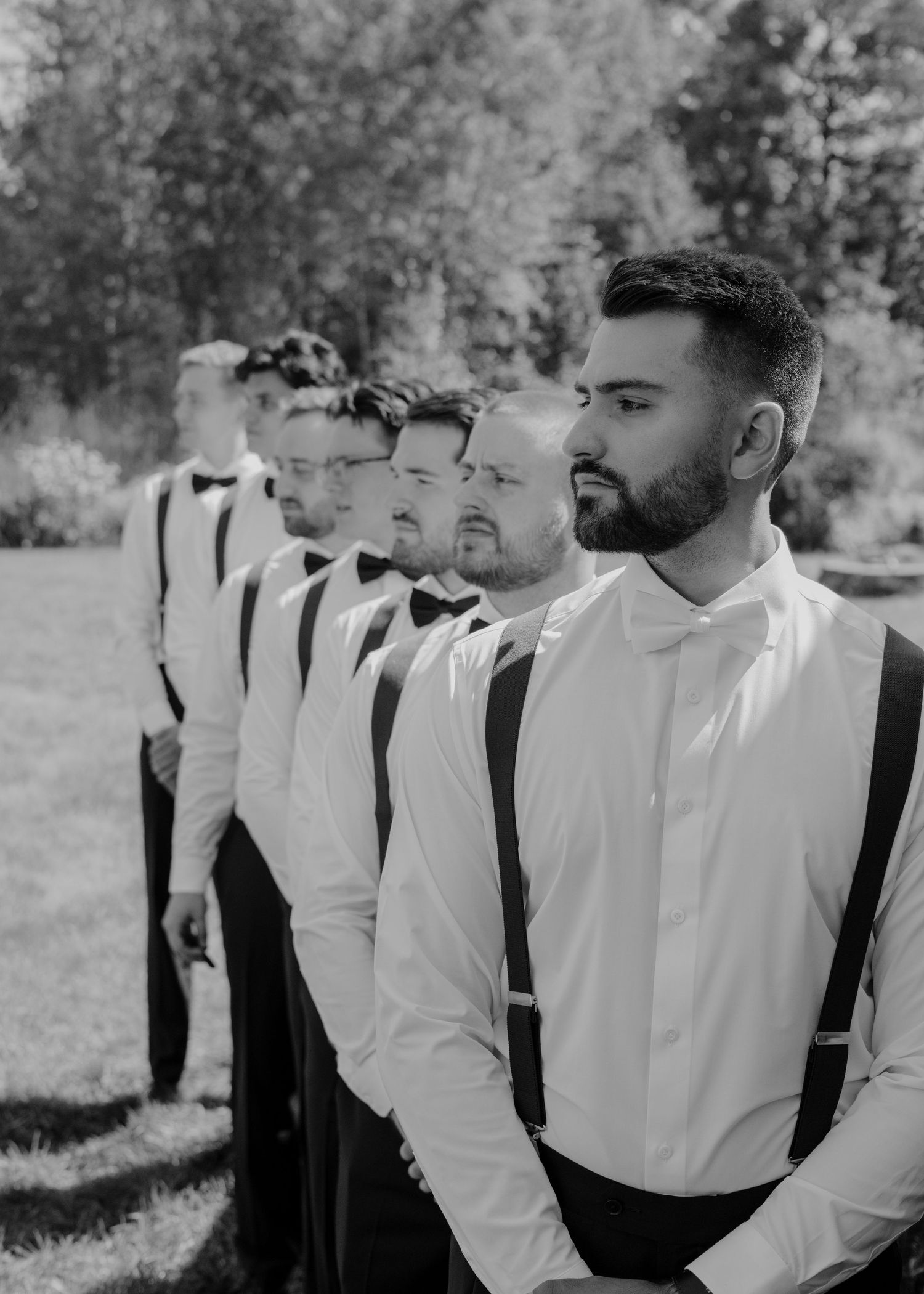 Groomsmen in white dress shirts and black suspenders stand in a row outdoors.