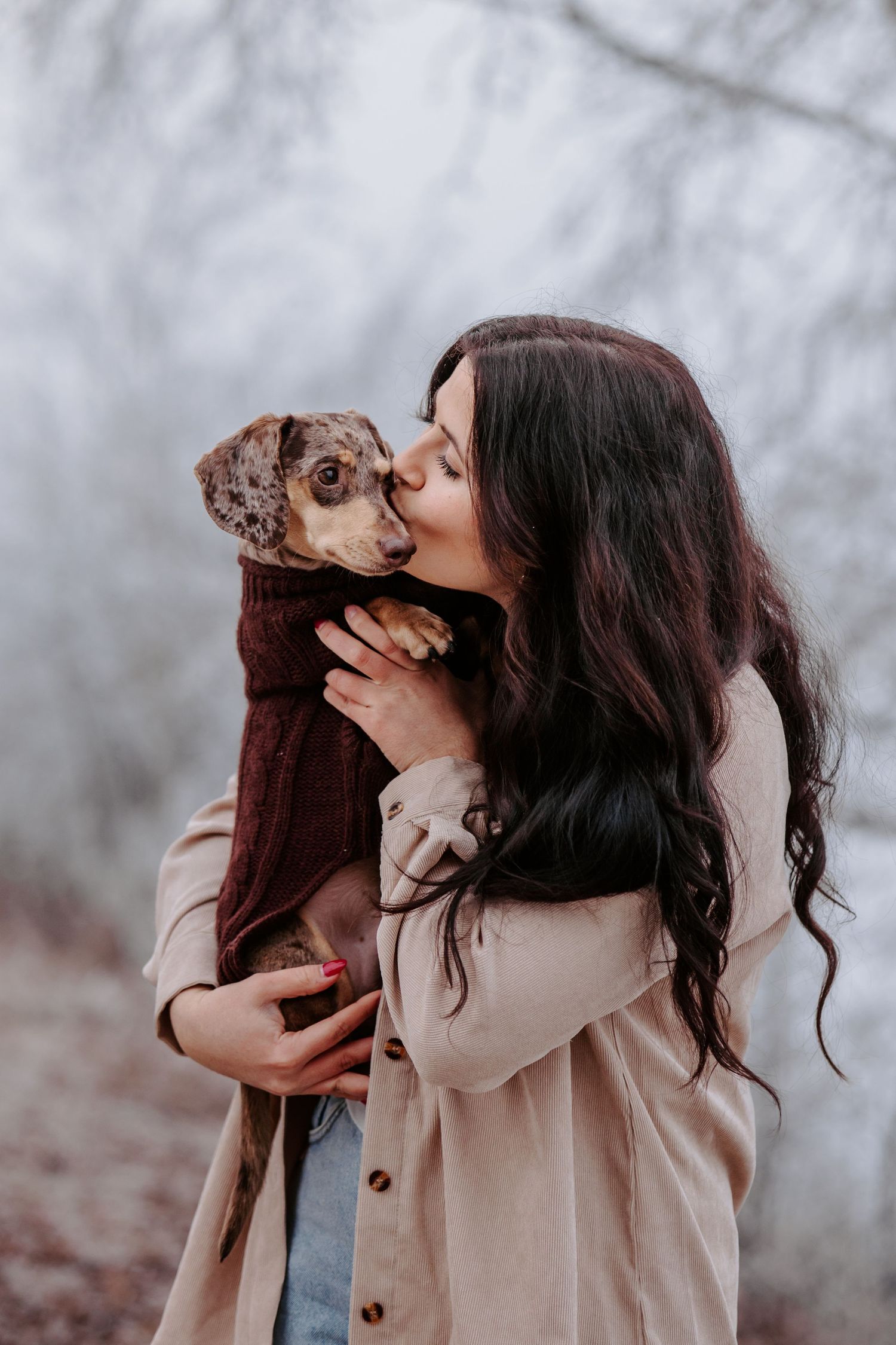 A woman in a beige coat kisses a small brown dachshund.