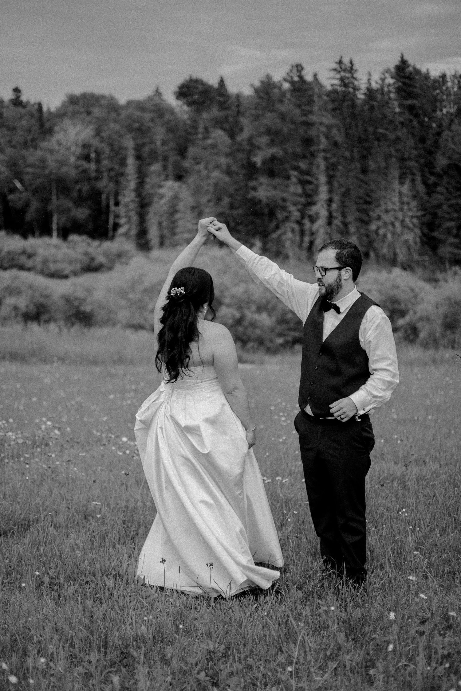 A couple dancing together in a field with evergreen trees in the background.