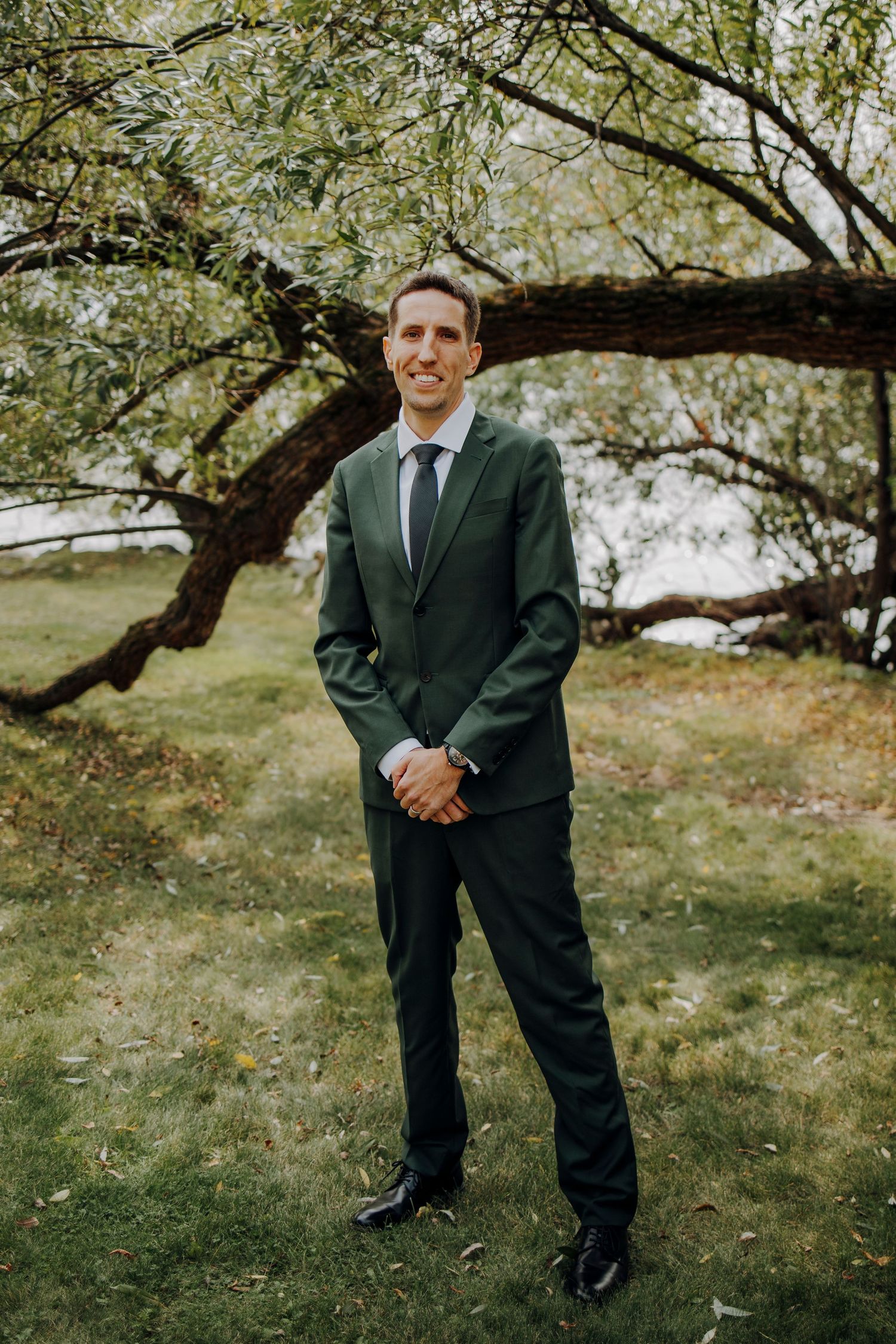 Groom in dark green suit poses casually under tree branches during outdoor wedding.