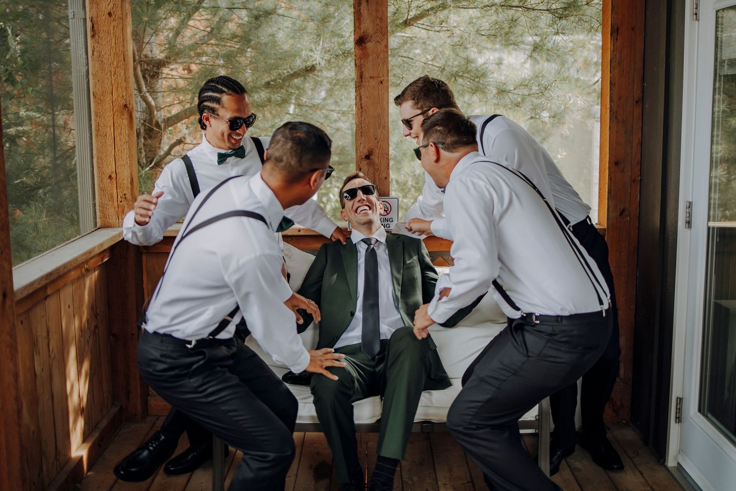 Groomsmen in suspenders and bow ties toast with drinks in a rustic wooden setting.