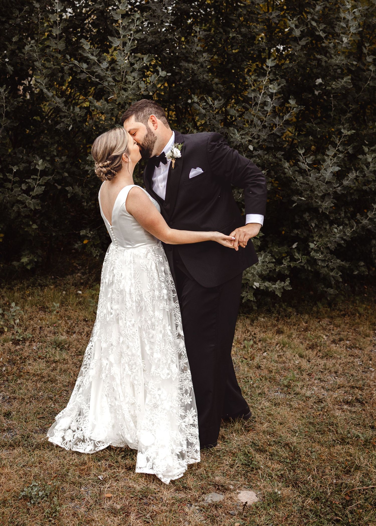 A romantic wedding couple sharing a kiss outdoors in front of foliage wearing a lace gown and black tuxedo.