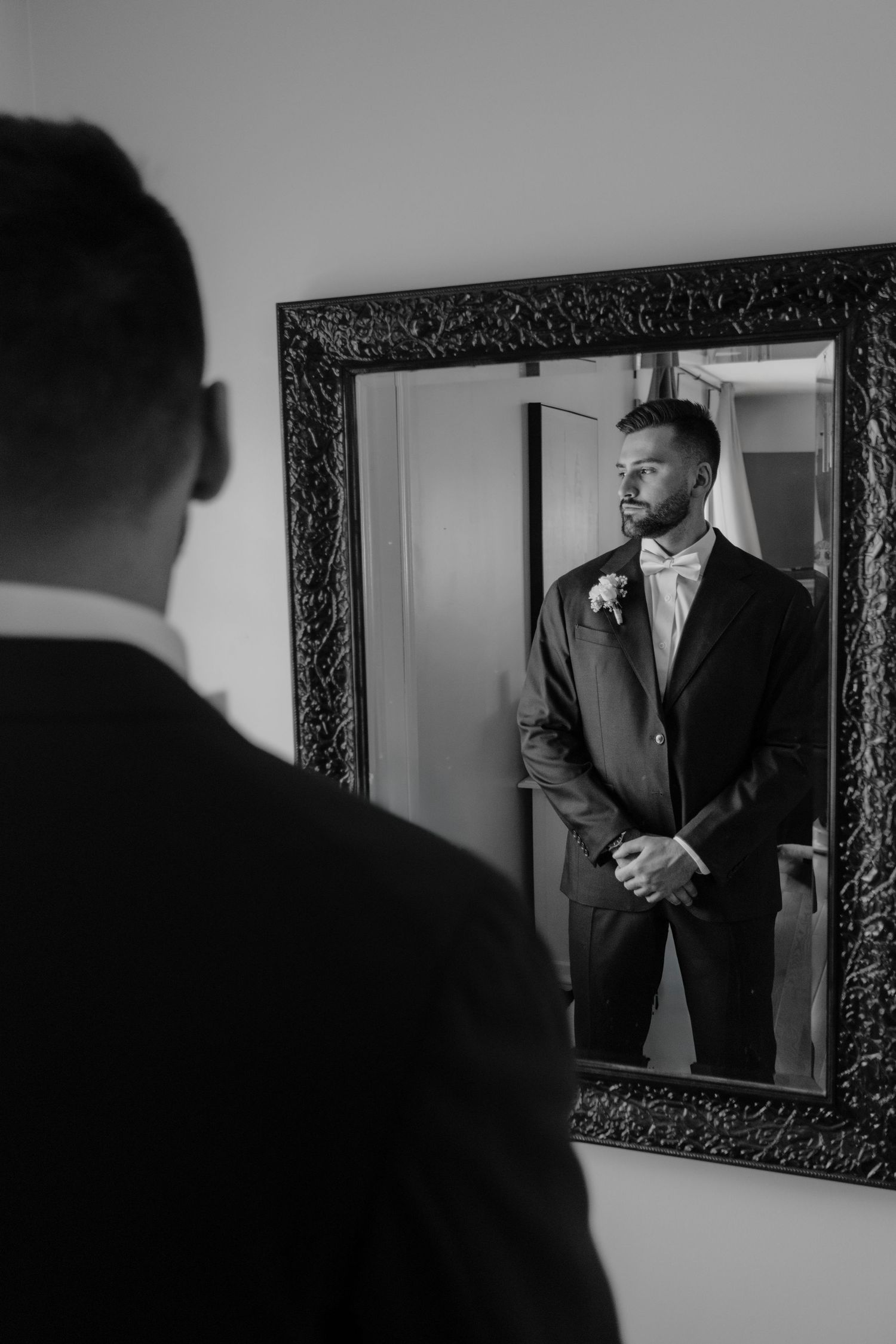 Black and white photo of a groom looking at his reflection in an ornate framed mirror.