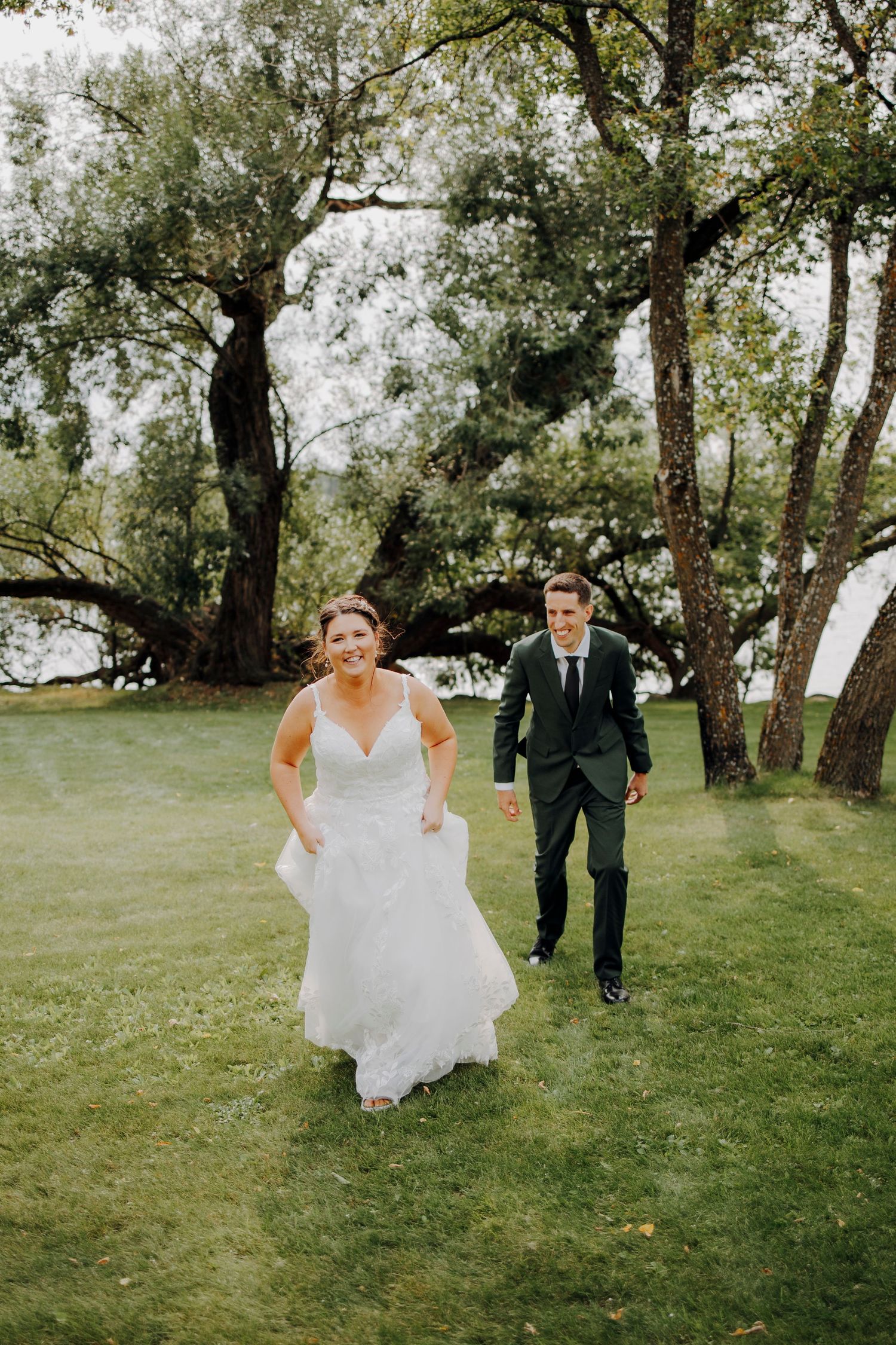 Wedding couple walking and playing in a grassy field surrounded by trees.