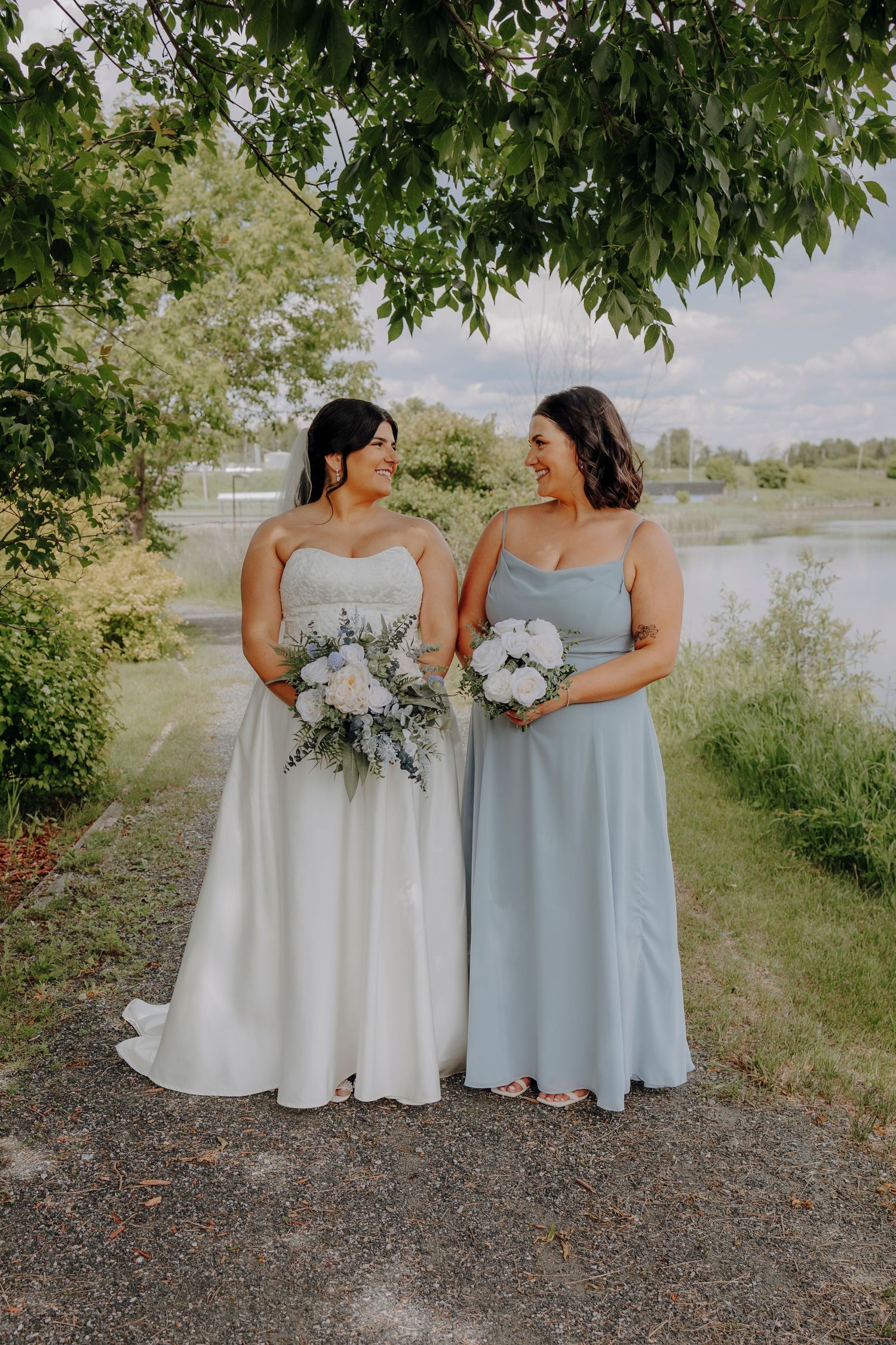 Two people in elegant dresses hold bouquets while standing on gravel path near waterfront.