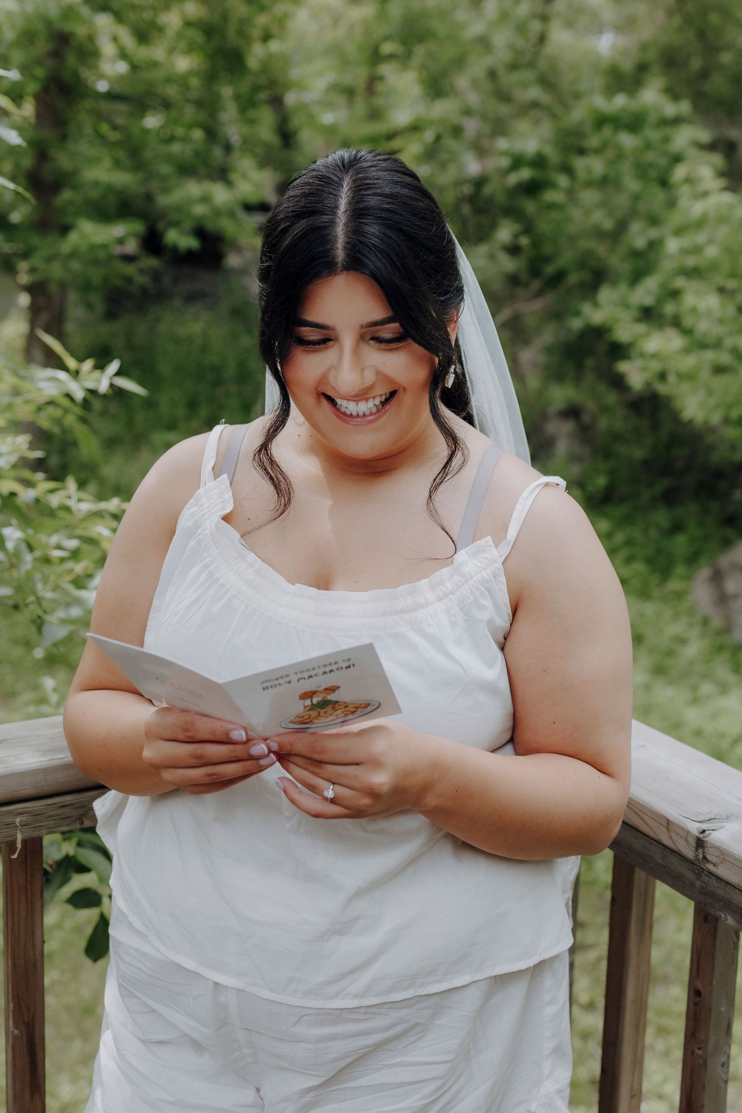A bride in a white dress sits on a wooden deck reading a letter with a joyful expression in an outdoor setting.