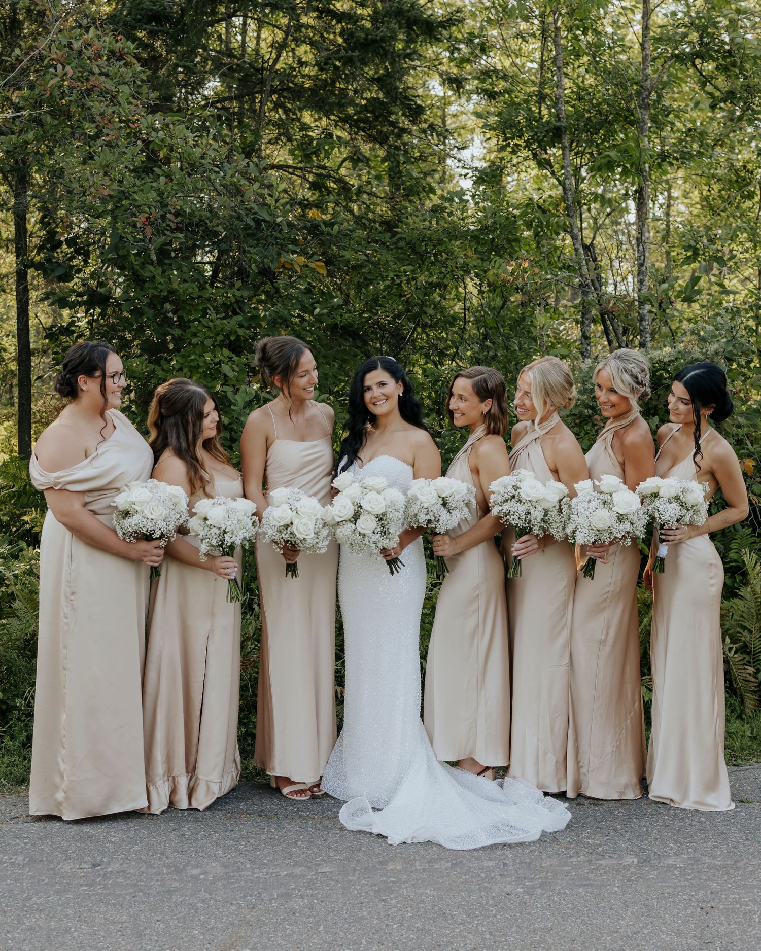 Bridesmaids in champagne dresses hold white bouquets while standing in a curved line outdoors.