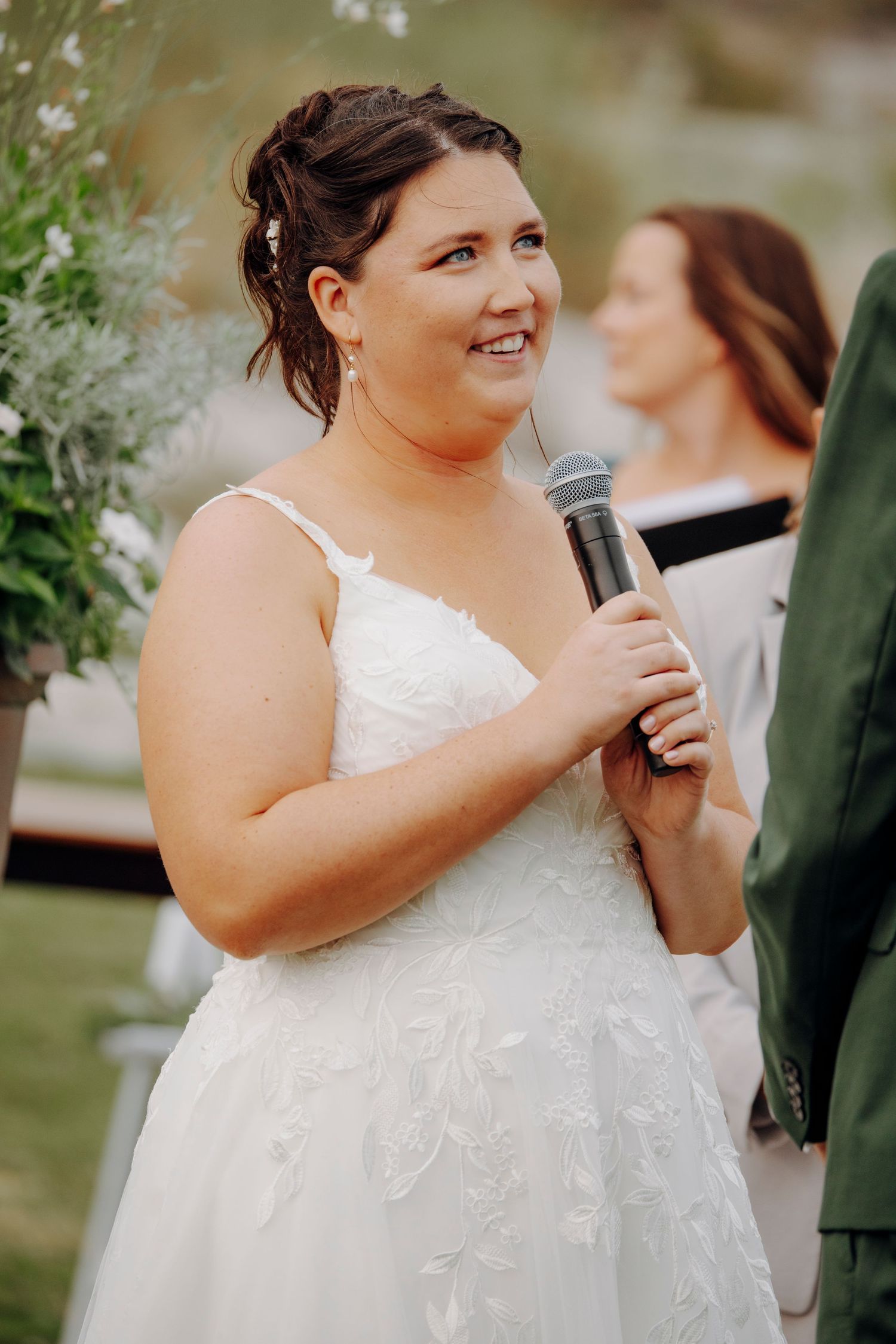 Bride in a white lace dress speaks into microphone during outdoor wedding ceremony.
