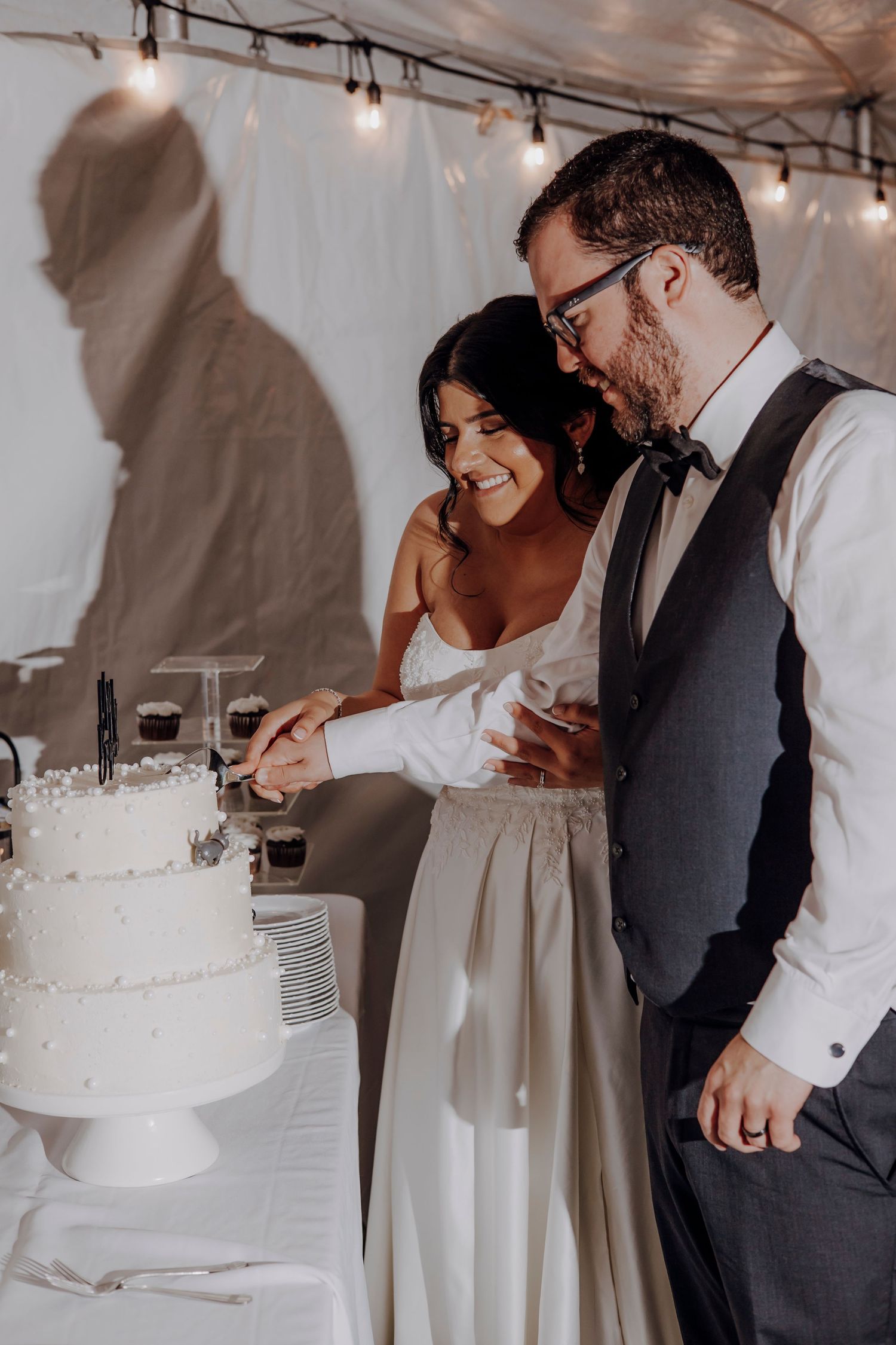 Wedding couple cuts their white tiered cake together under string lights at reception.