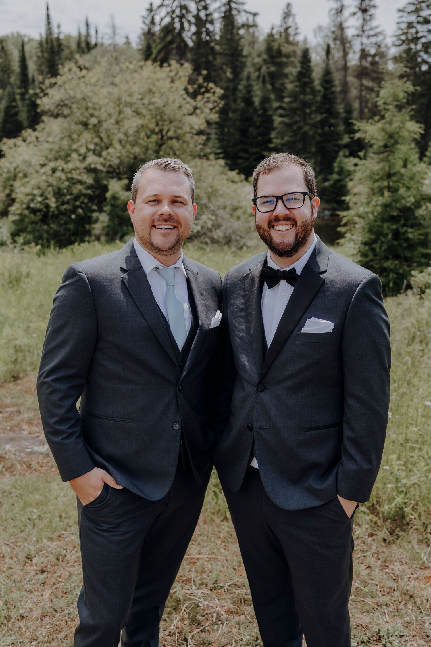 Two people in black suits and bowties stand together in a forest setting with evergreen trees in the background.