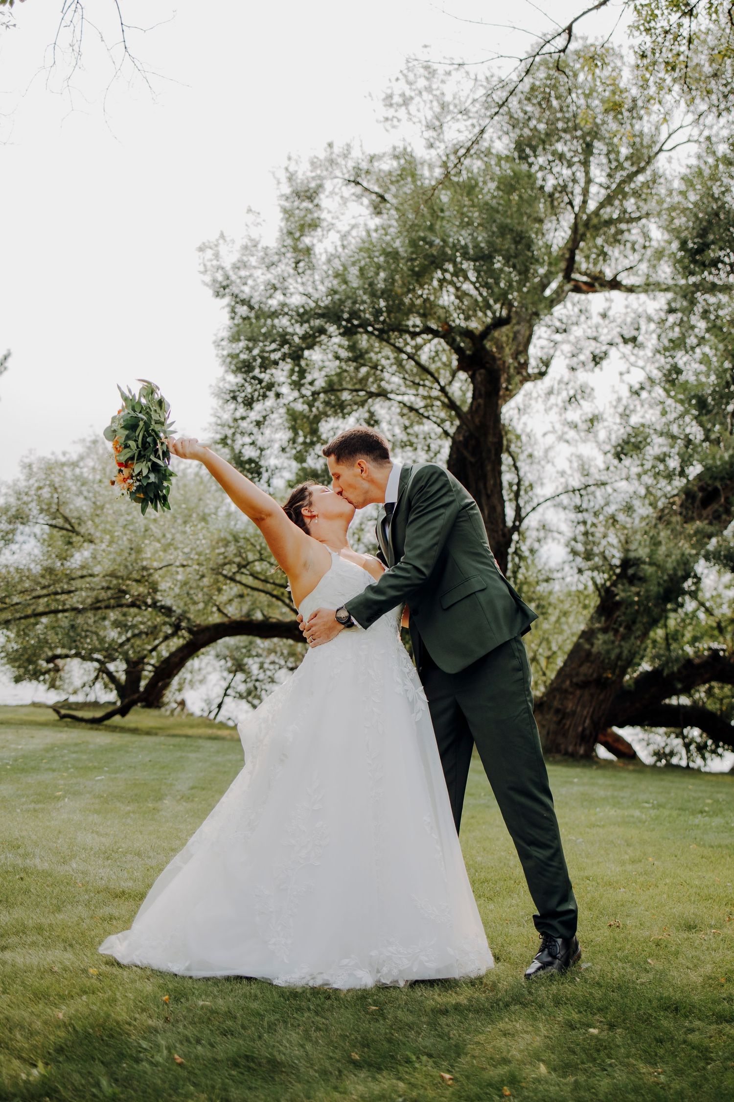 A couple performs a romantic dip dance pose on grassy grounds with trees in the background.
