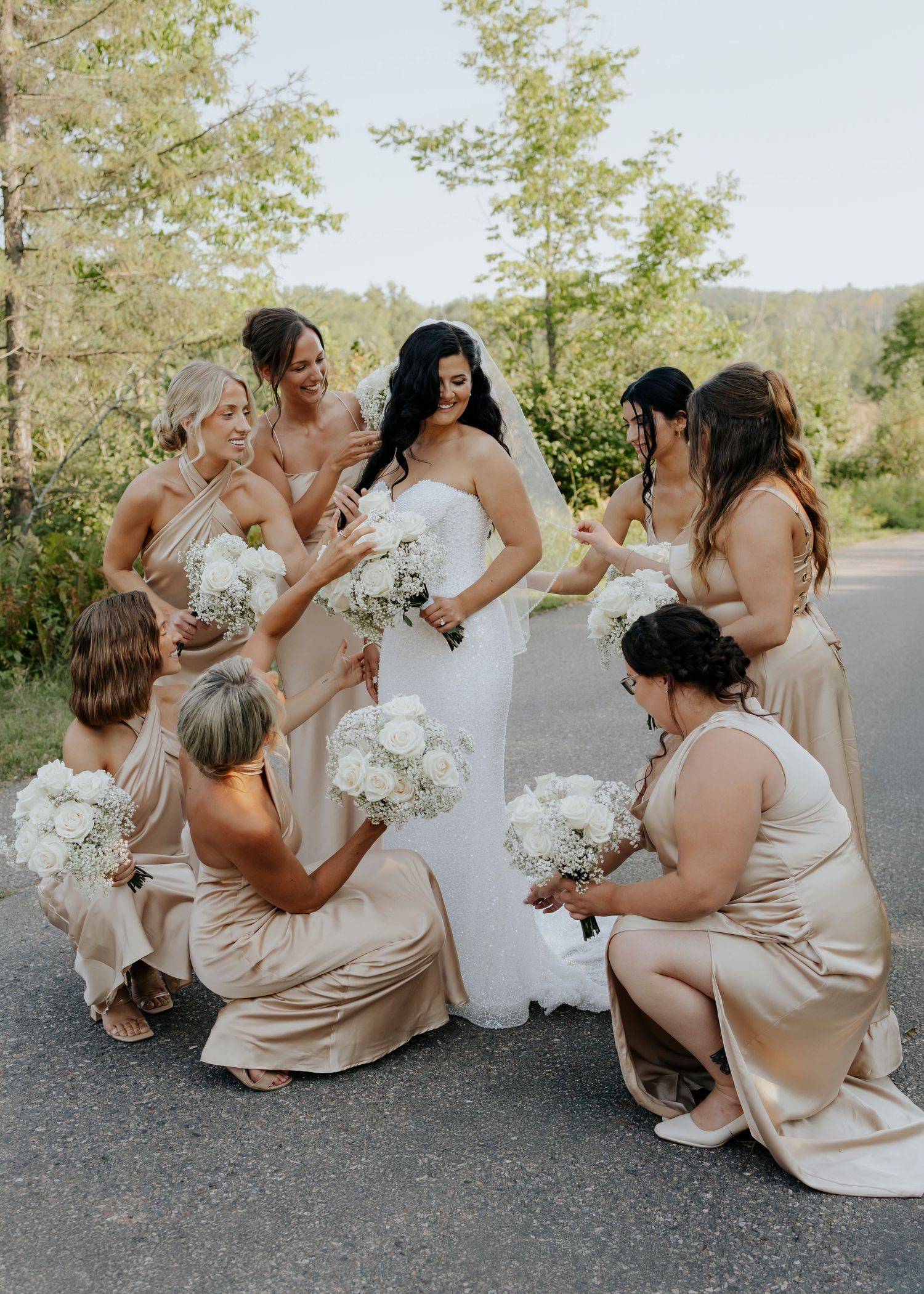 Bridesmaids in champagne dresses gather around the bride's dress outdoors.