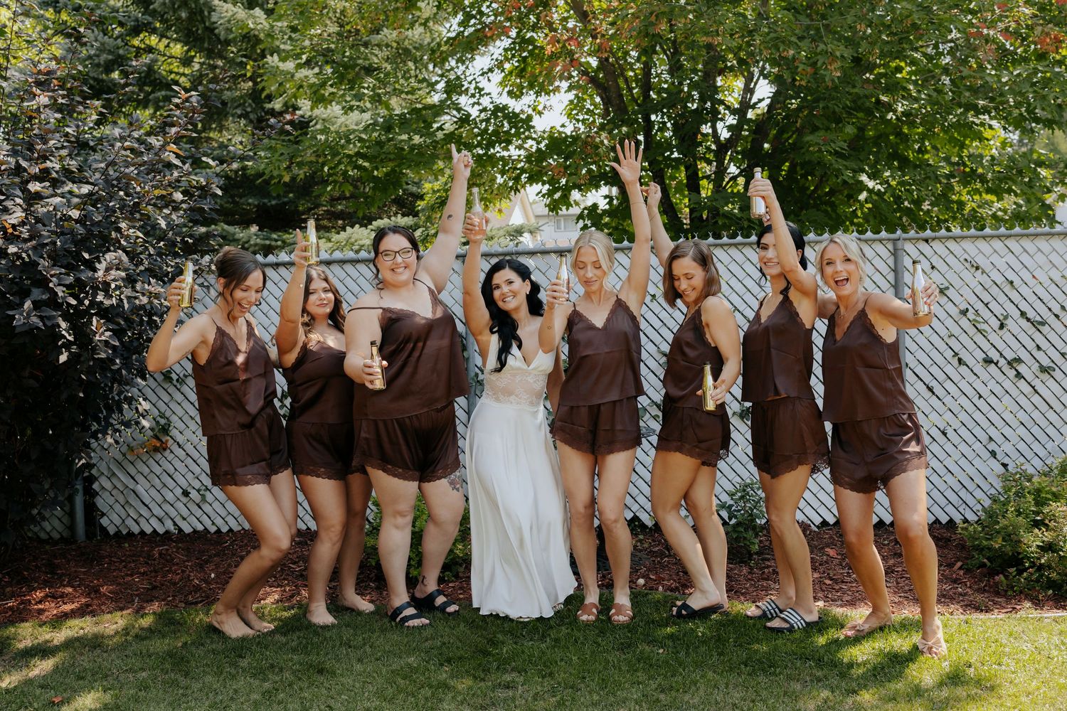 Wedding party showing off their brown shoes lined up against white fence in outdoor setting.
