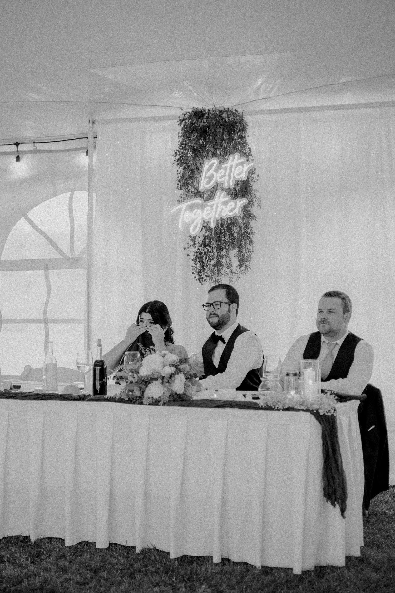 A black and white photo of wedding party members seated at a head table with floral decorations.