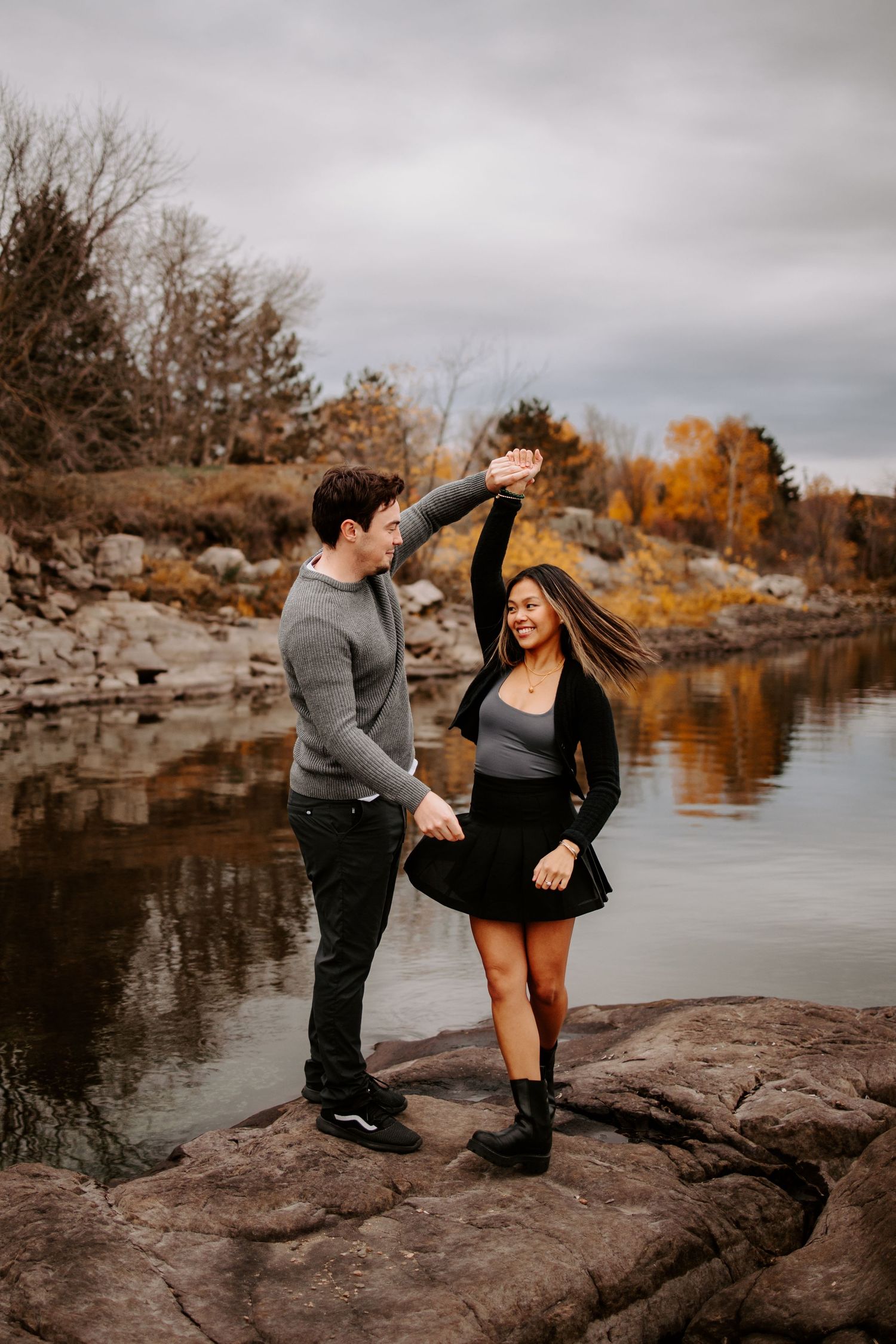 A couple dances on rocky shore with autumn trees and reflective water in the background.