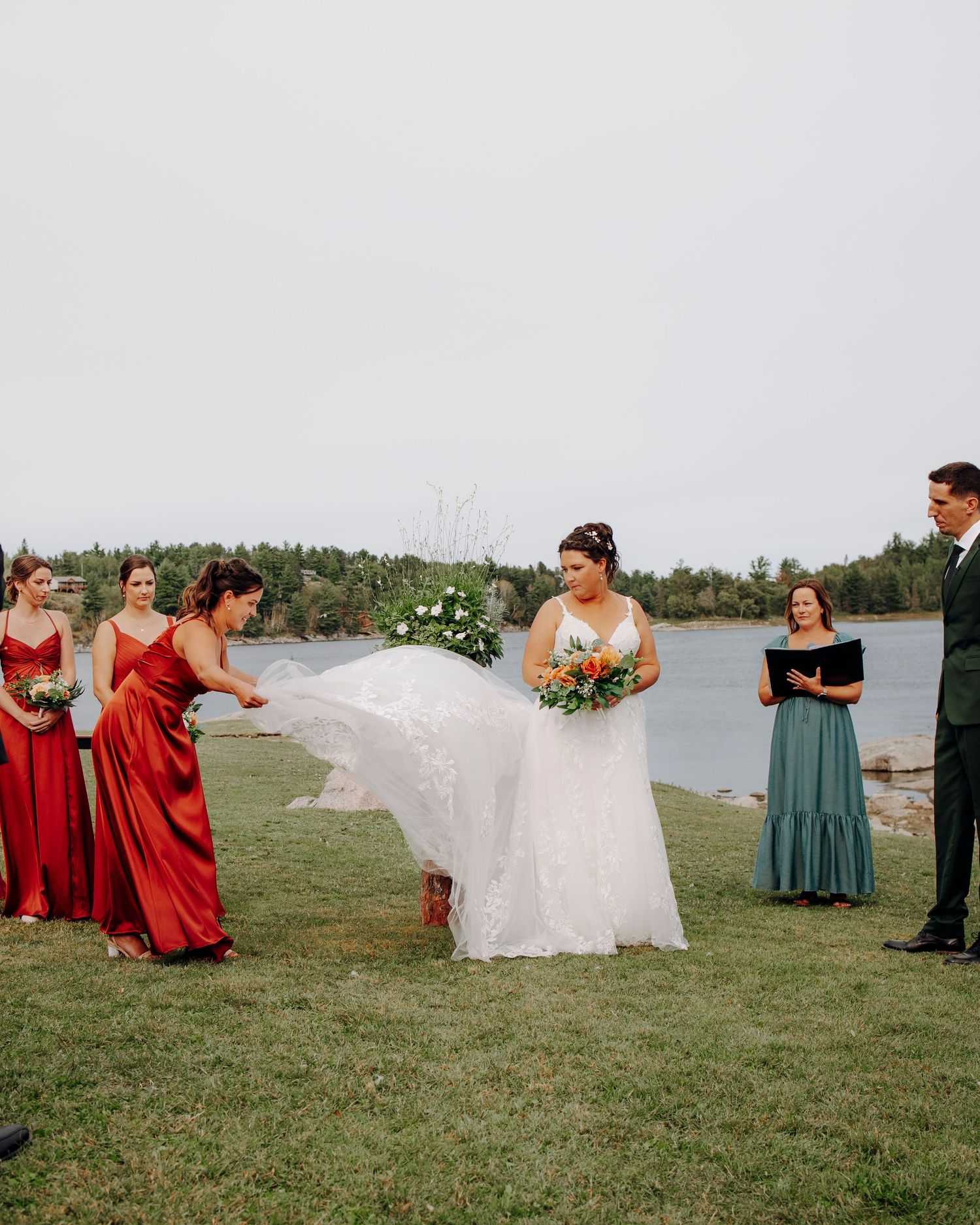 Wedding party posing playfully with flowing white dress on a lakeside lawn with bridesmaids in red and teal.
