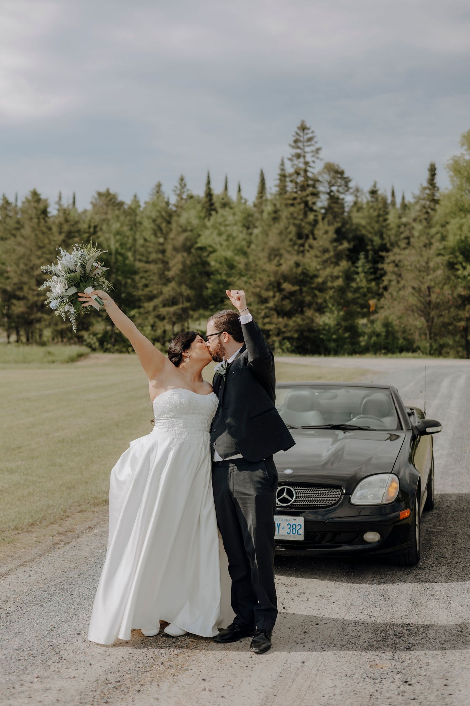 A couple in wedding attire poses playfully near a black Mercedes-Benz on a gravel road with evergreen trees in background.