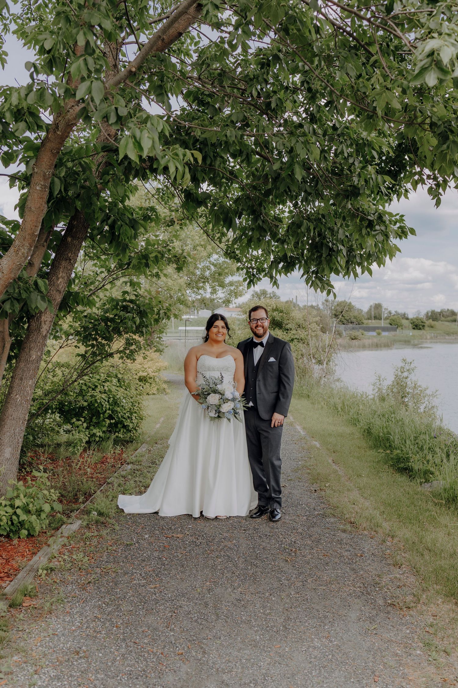 Newlyweds walk together on a scenic tree-lined path near a lake.