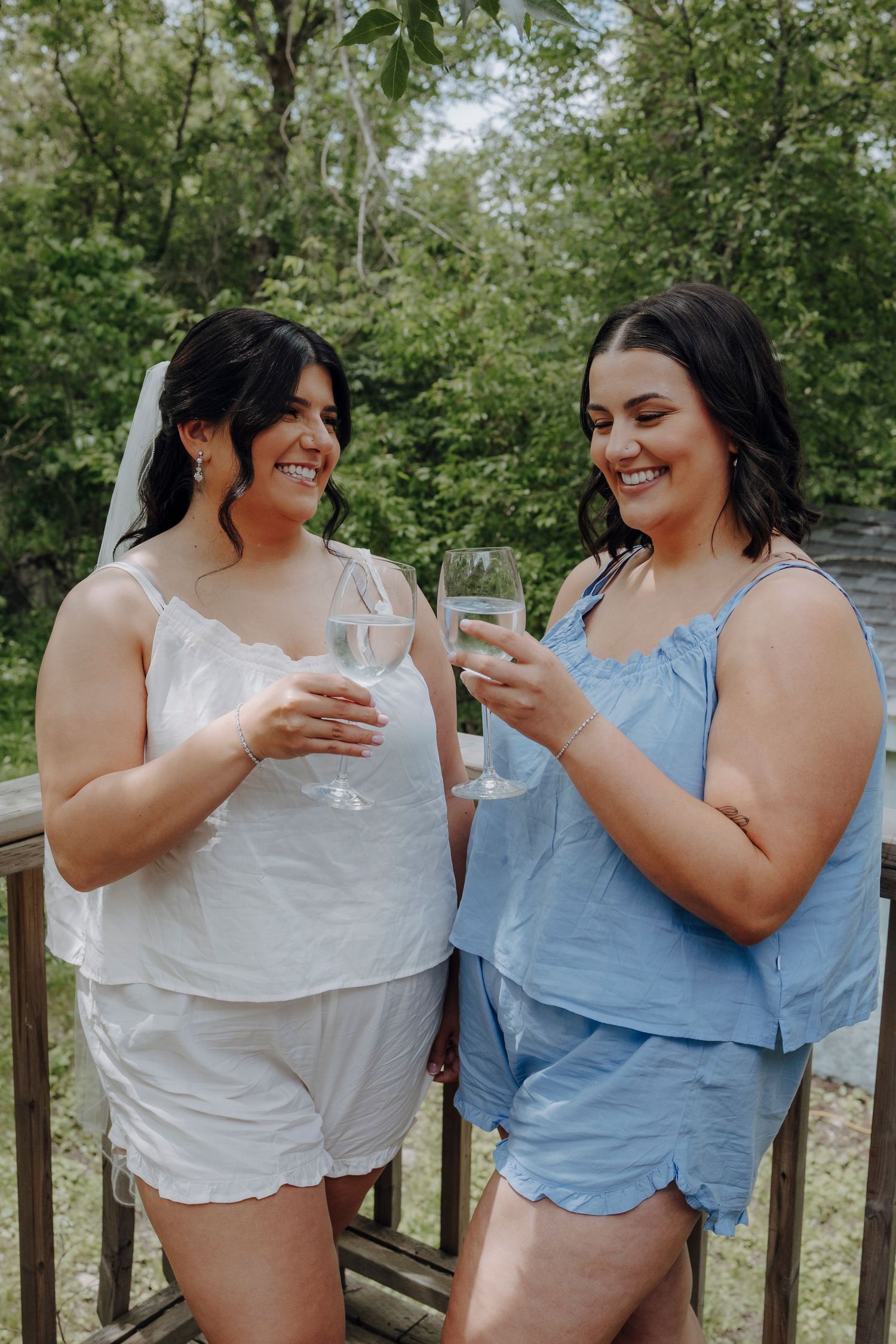 Two friends in white and blue summer outfits share laughter while holding drinks on a wooden deck outdoors.