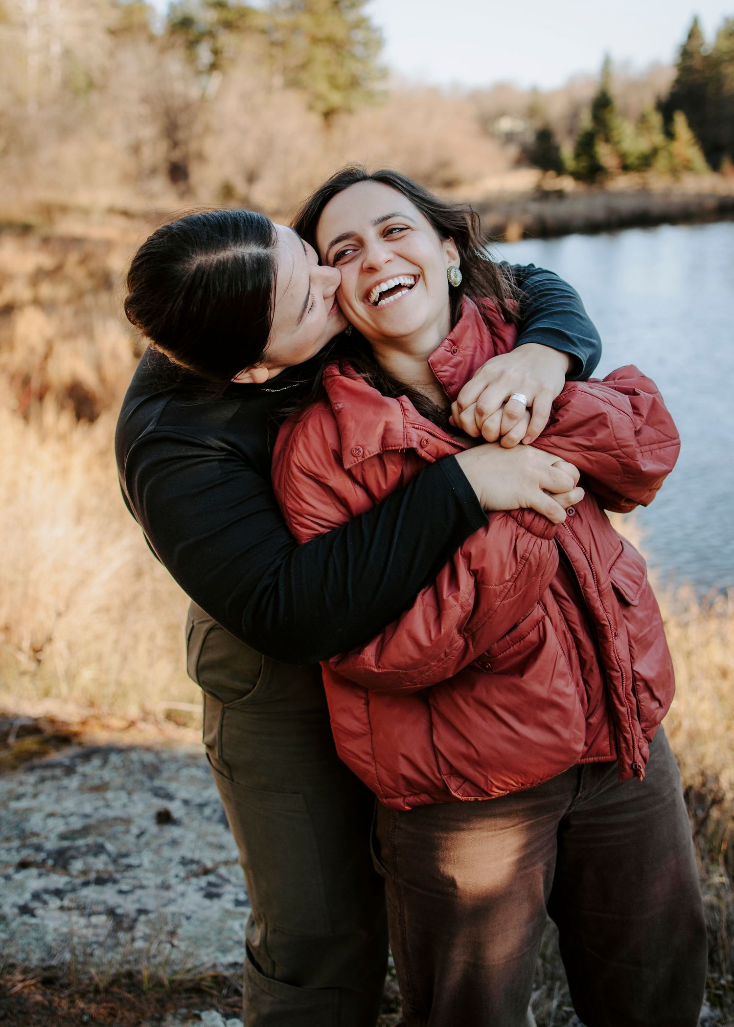 Two people sharing a joyful embrace and laughing together by a lake on a sunny autumn day.