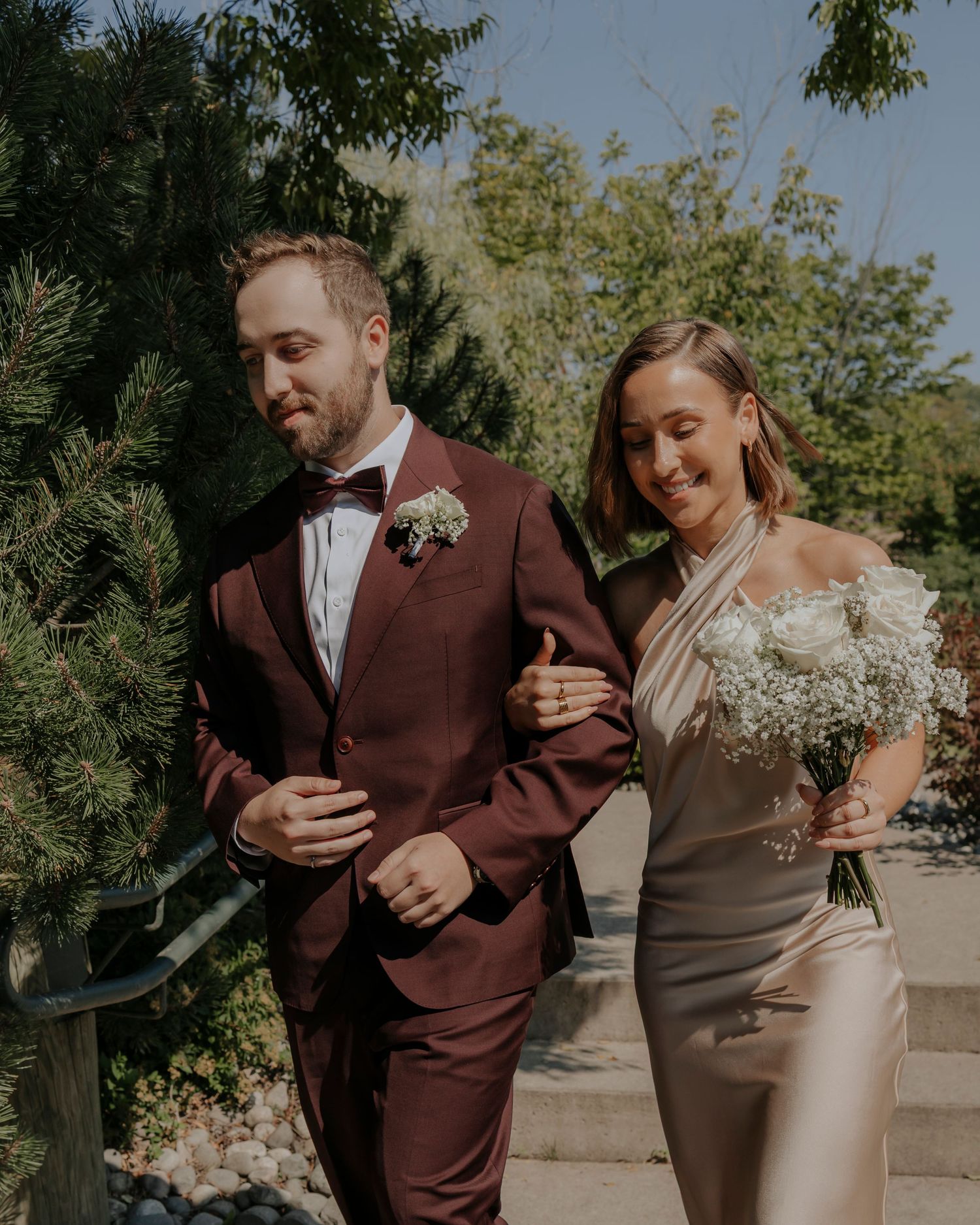 A couple in wedding attire, with the groom wearing a brown suit and bride in a fitted champagne dress walking outdoors.
