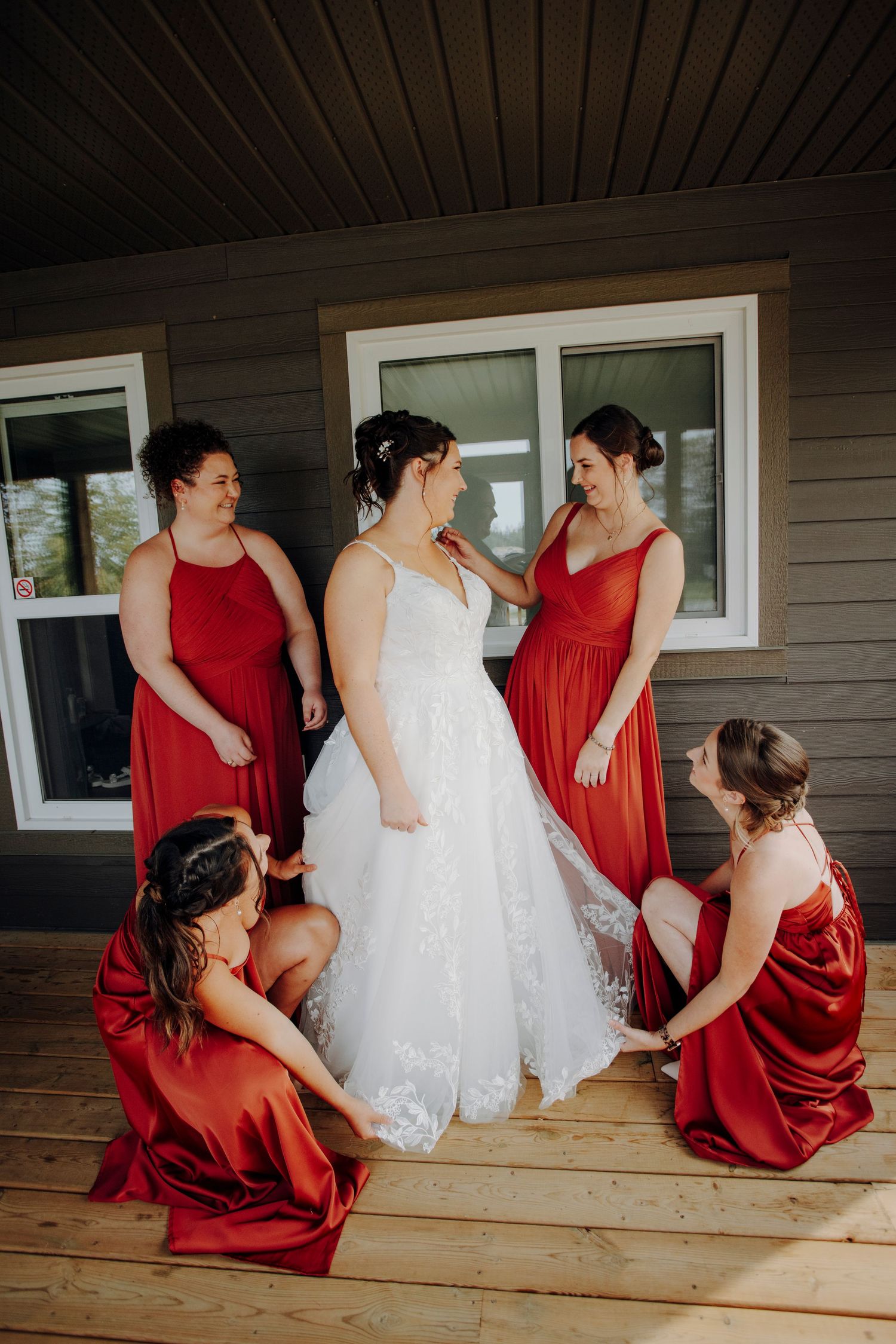 Wedding party gathered on a wooden deck with bridesmaids in red dresses helping with the bride's white gown.