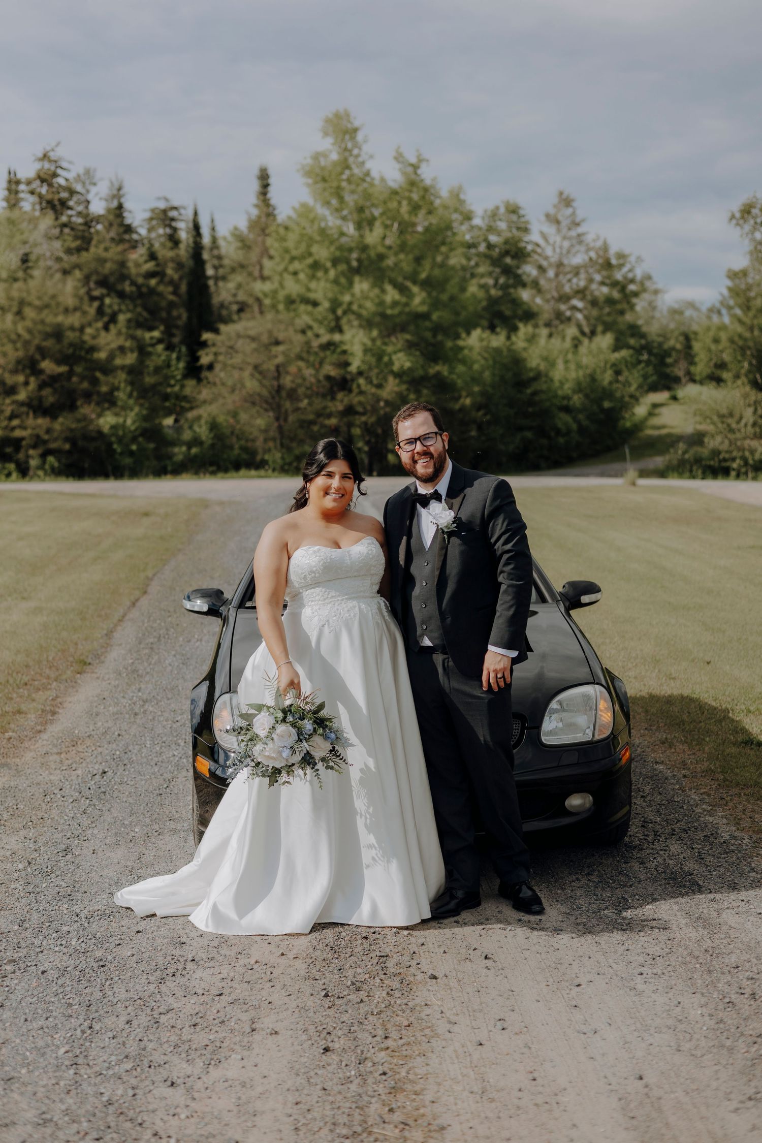 Wedding couple poses casually next to black car on gravel road surrounded by green landscape.