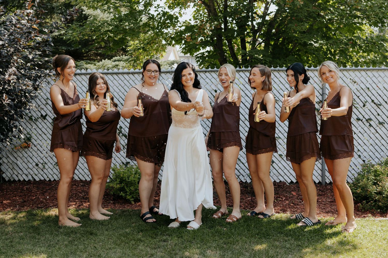 A wedding party wearing coordinating brown dresses poses together in a backyard garden setting.