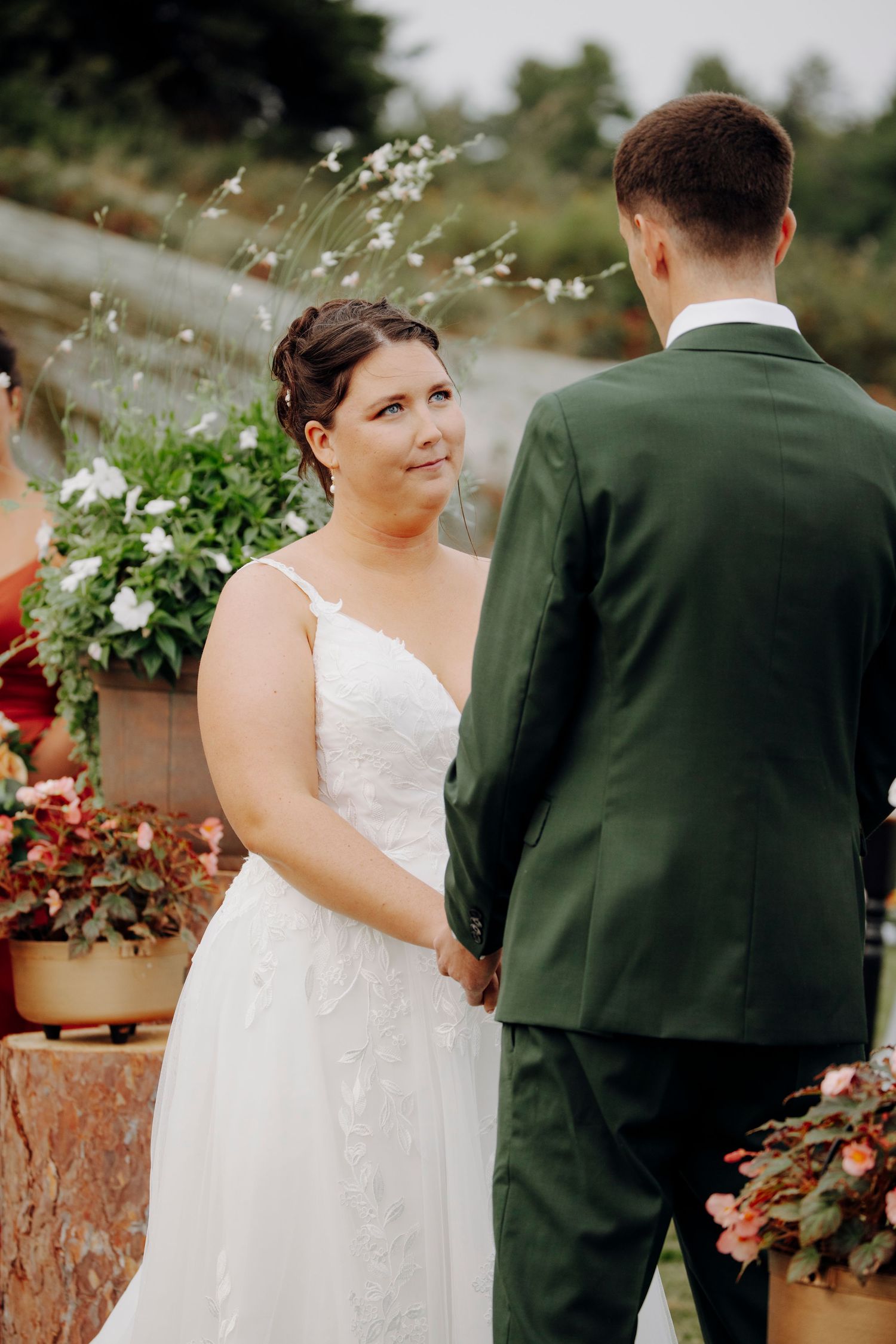 Outdoor wedding ceremony with couple in white dress and dark suit standing together among greenery.