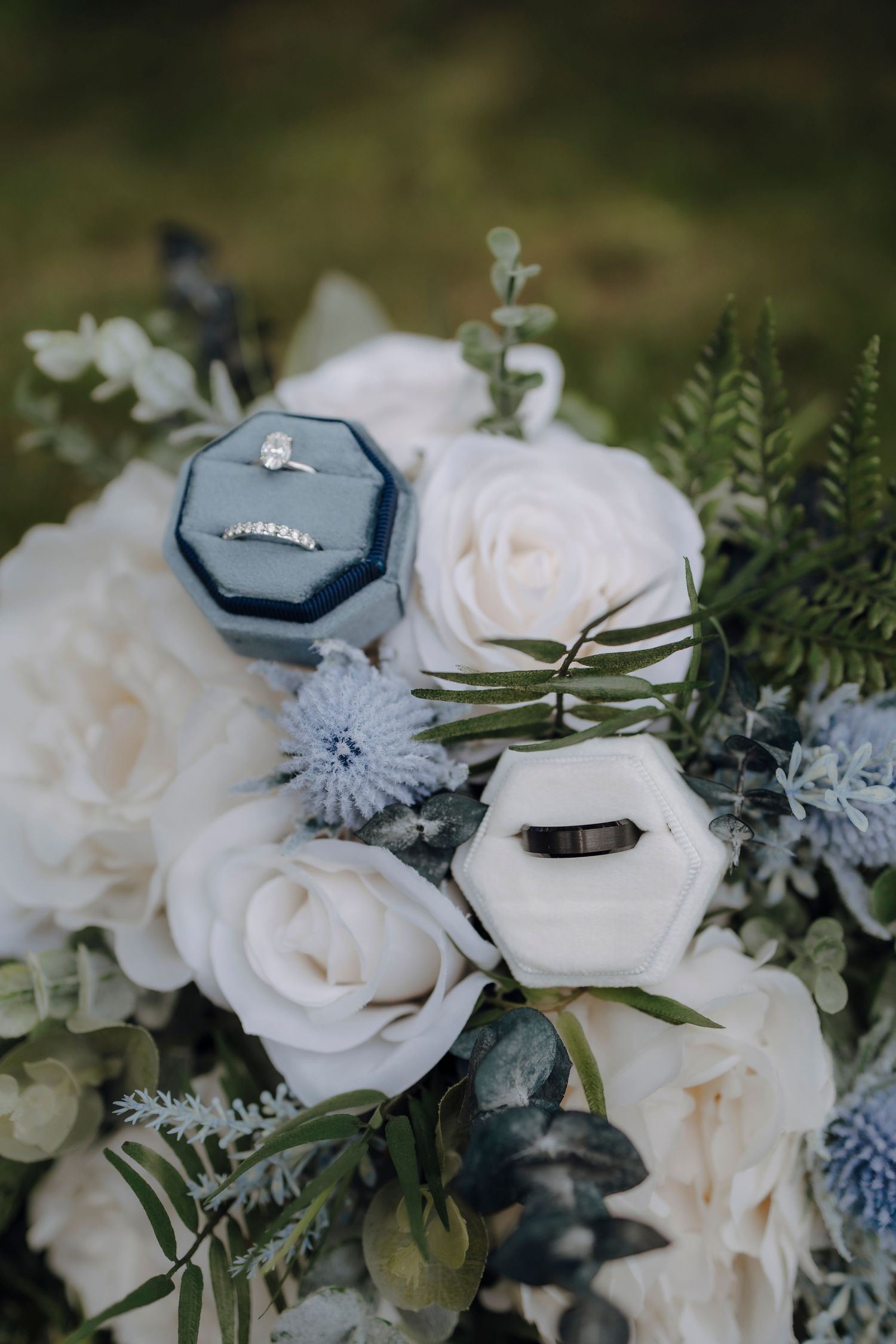 Wedding rings nestled in a dusty blue velvet box surrounded by white roses and delicate blue floral accents.