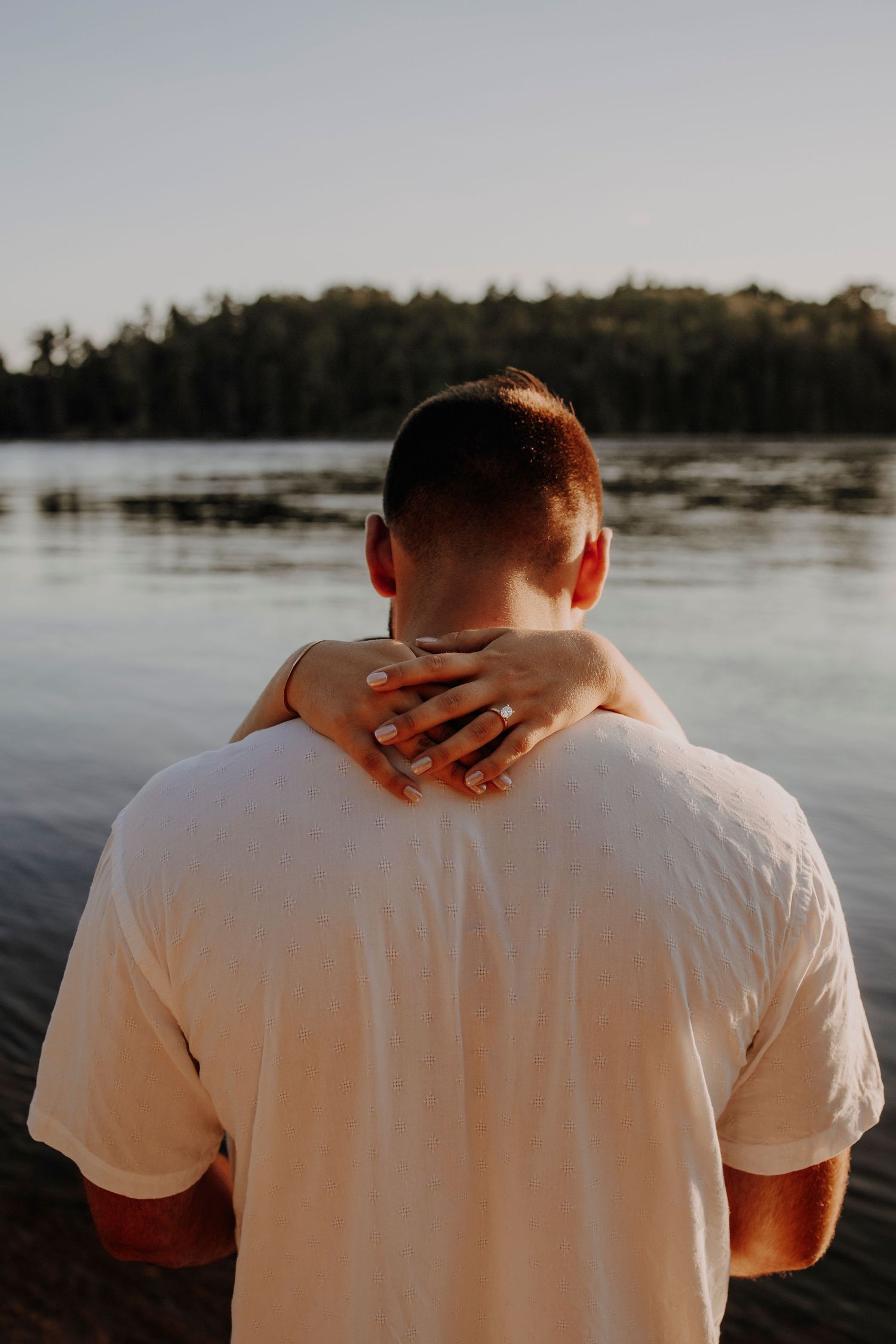 Person in white shirt being embraced from behind with hands on shoulders near a lake.