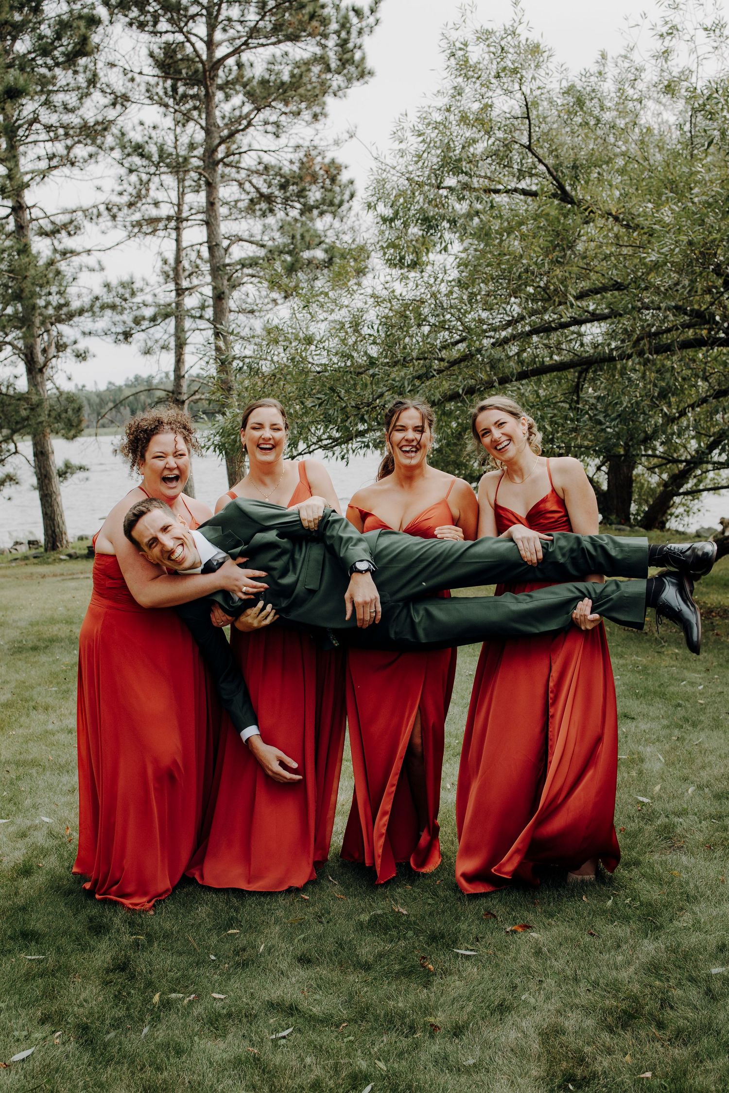 Bridesmaids in flowing red dresses pose playfully on grass for a group photo.
