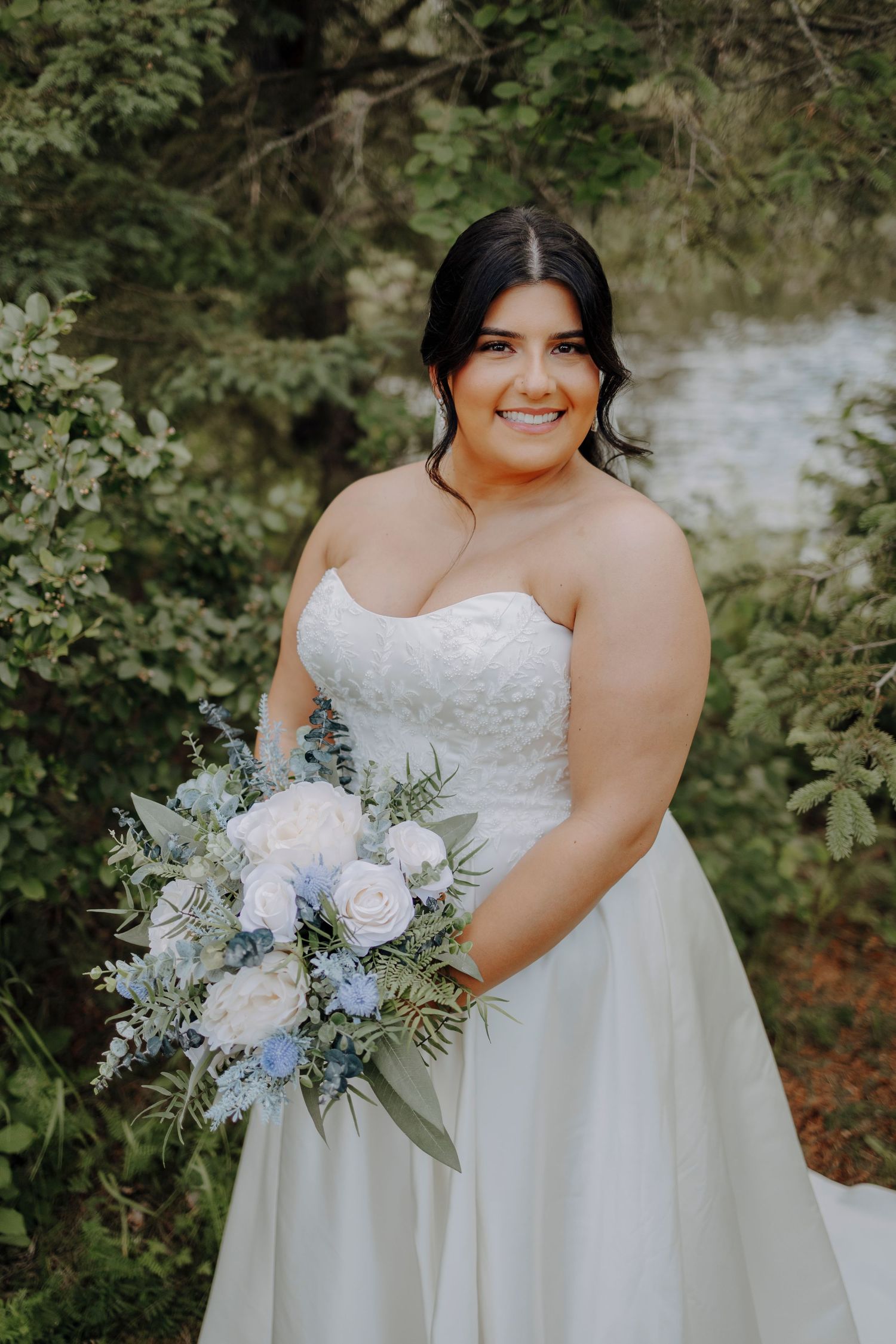 Bride in a strapless white wedding dress holds a blue and white floral bouquet against a natural outdoor backdrop.