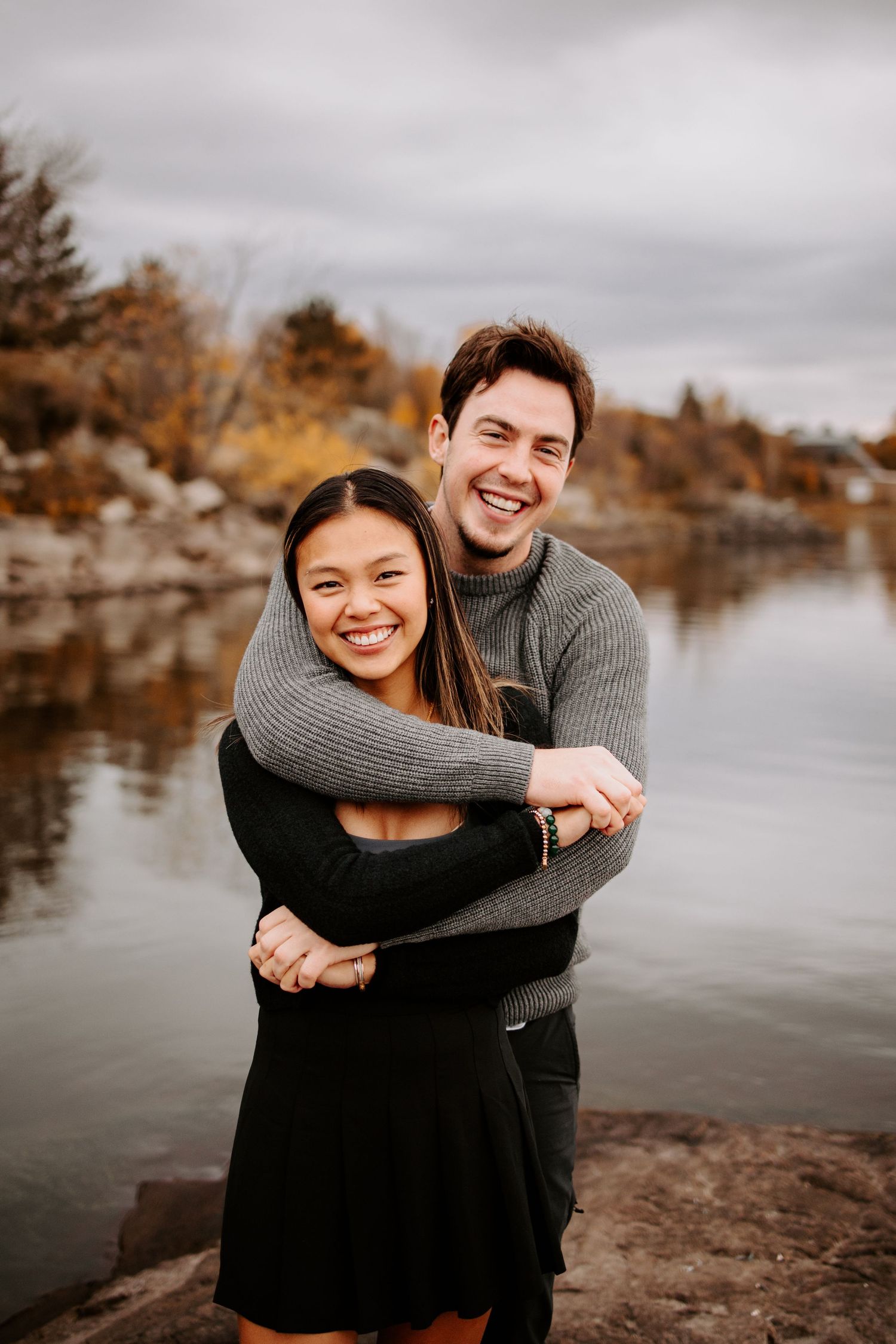 A couple shares a warm embrace while standing on rocks near a serene lake in autumn.