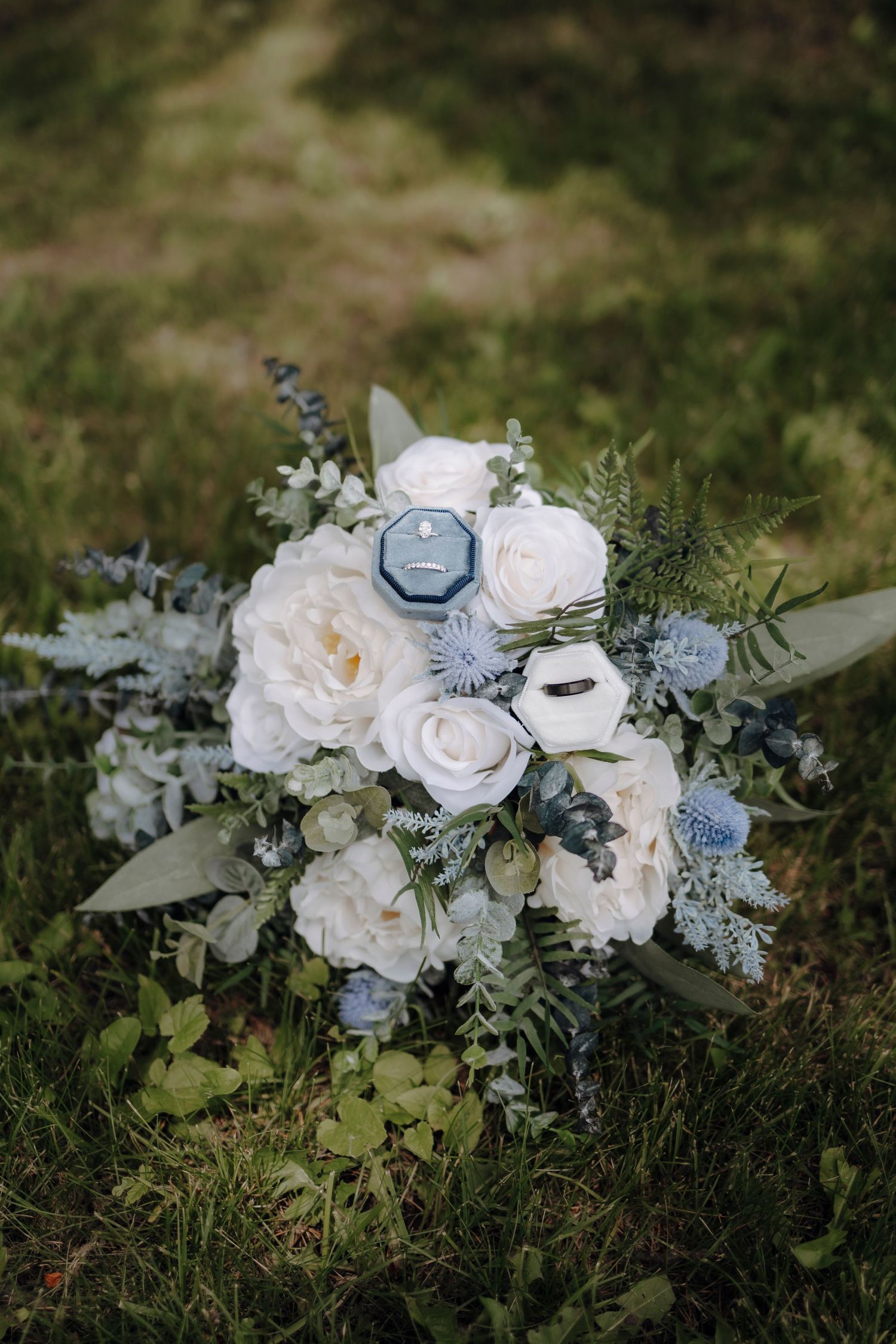 A romantic wedding bouquet with white roses, blue thistle, and delicate greenery arranged on grass in natural light.