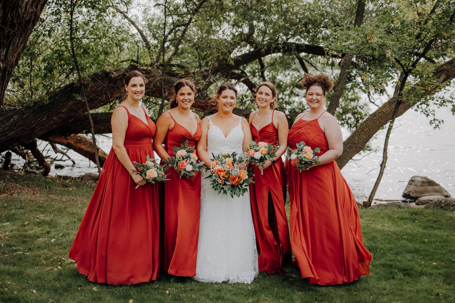 Bridal party in vibrant red dresses poses under tree branches with colorful bouquets.