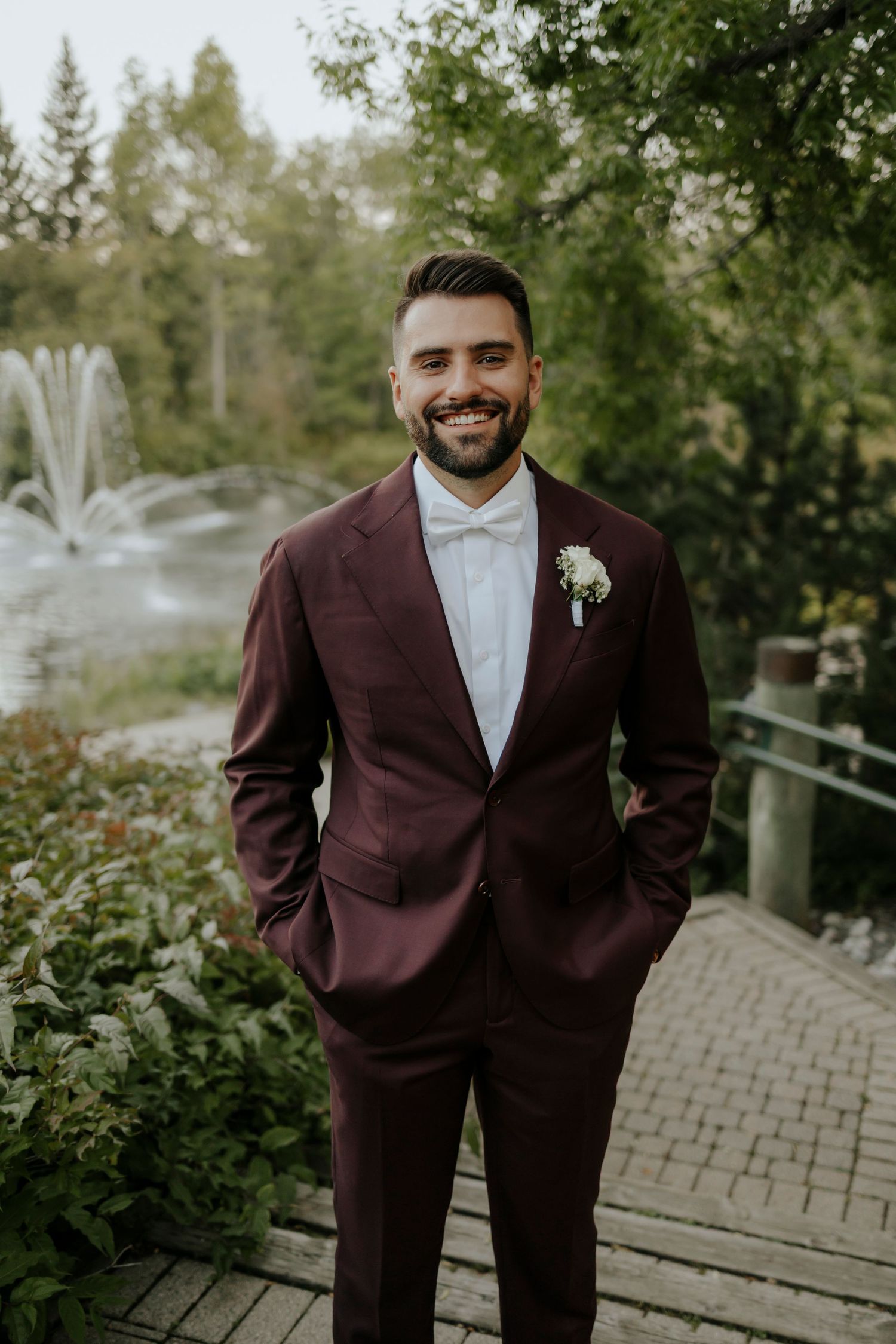 A person in a burgundy suit poses on wooden decking surrounded by greenery.