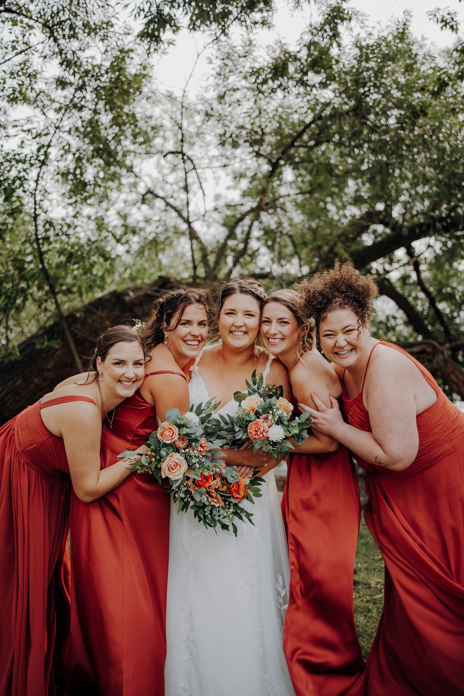 Wedding party in red bridesmaid dresses huddled together in a joyful group pose with floral bouquets outdoors.