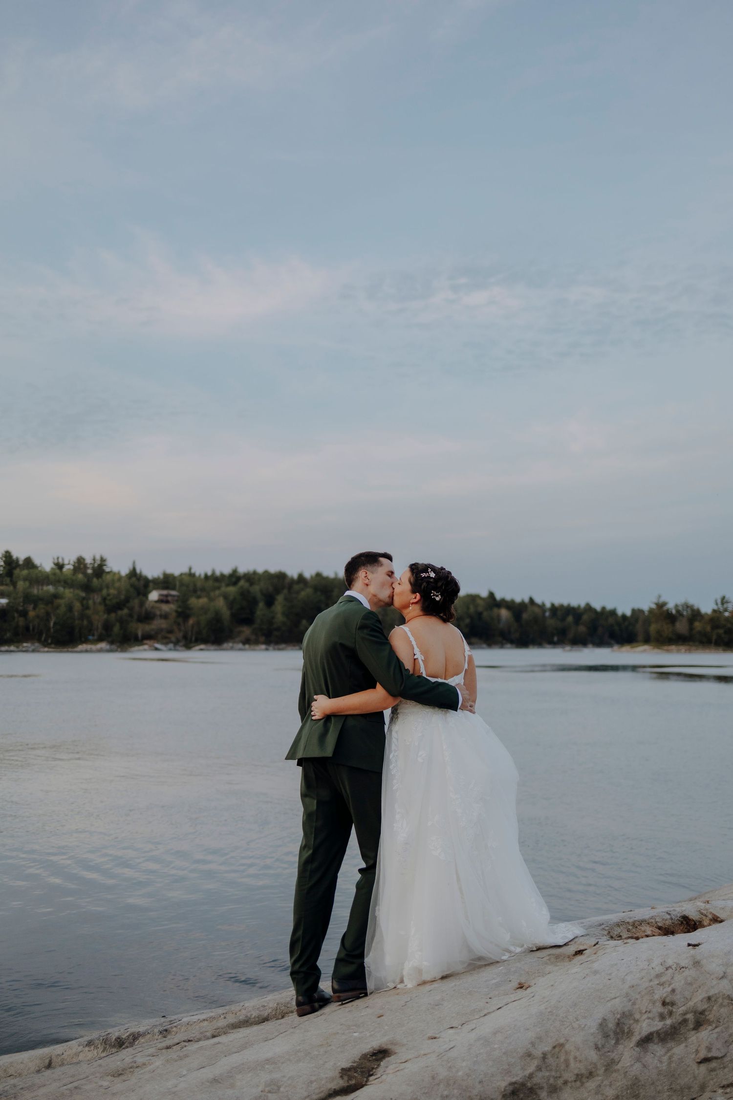 Newlyweds share an intimate moment on rocky outcrop overlooking a serene lake.
