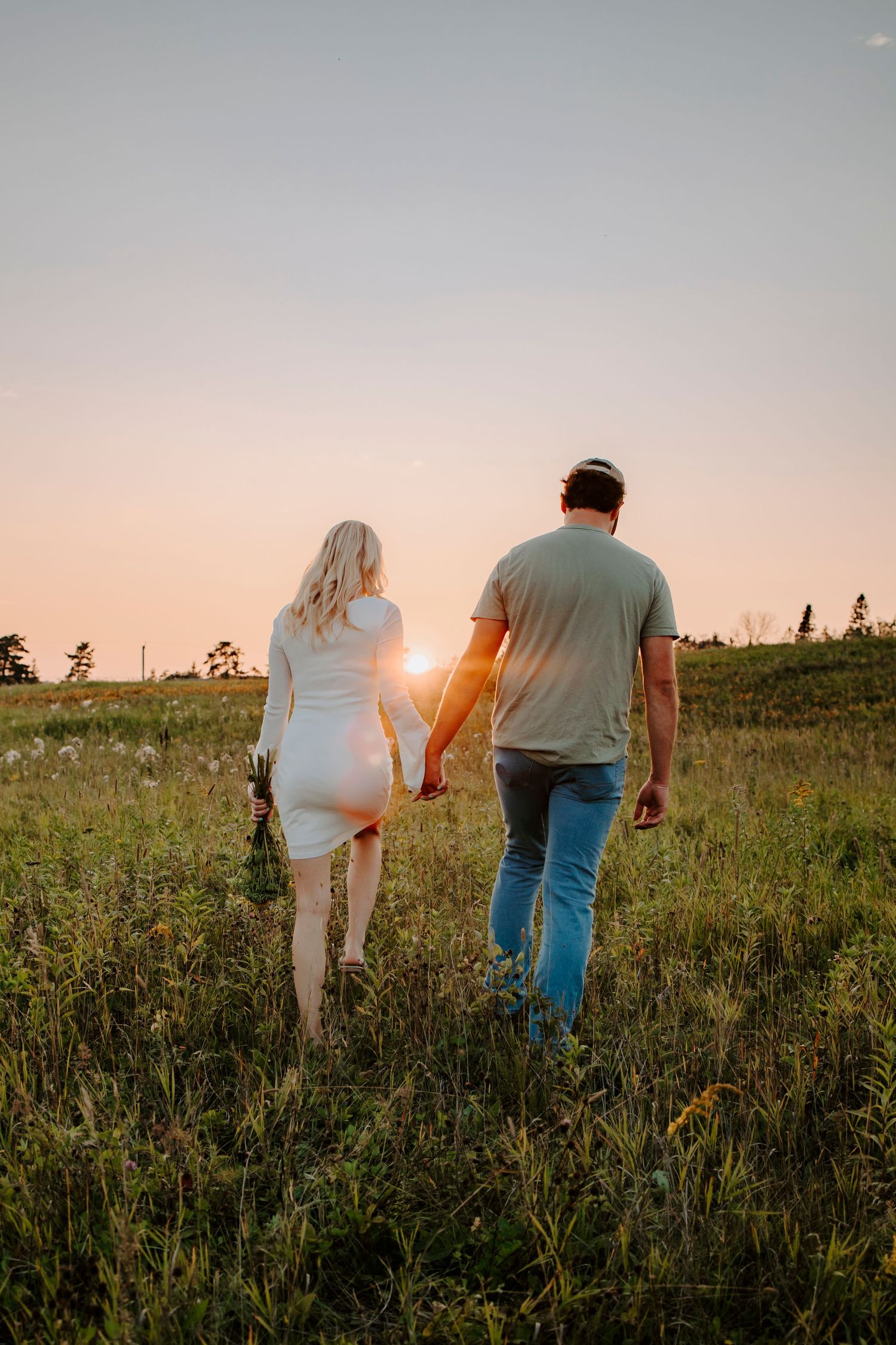 Couple walking through a grassy field at sunset holding hands.